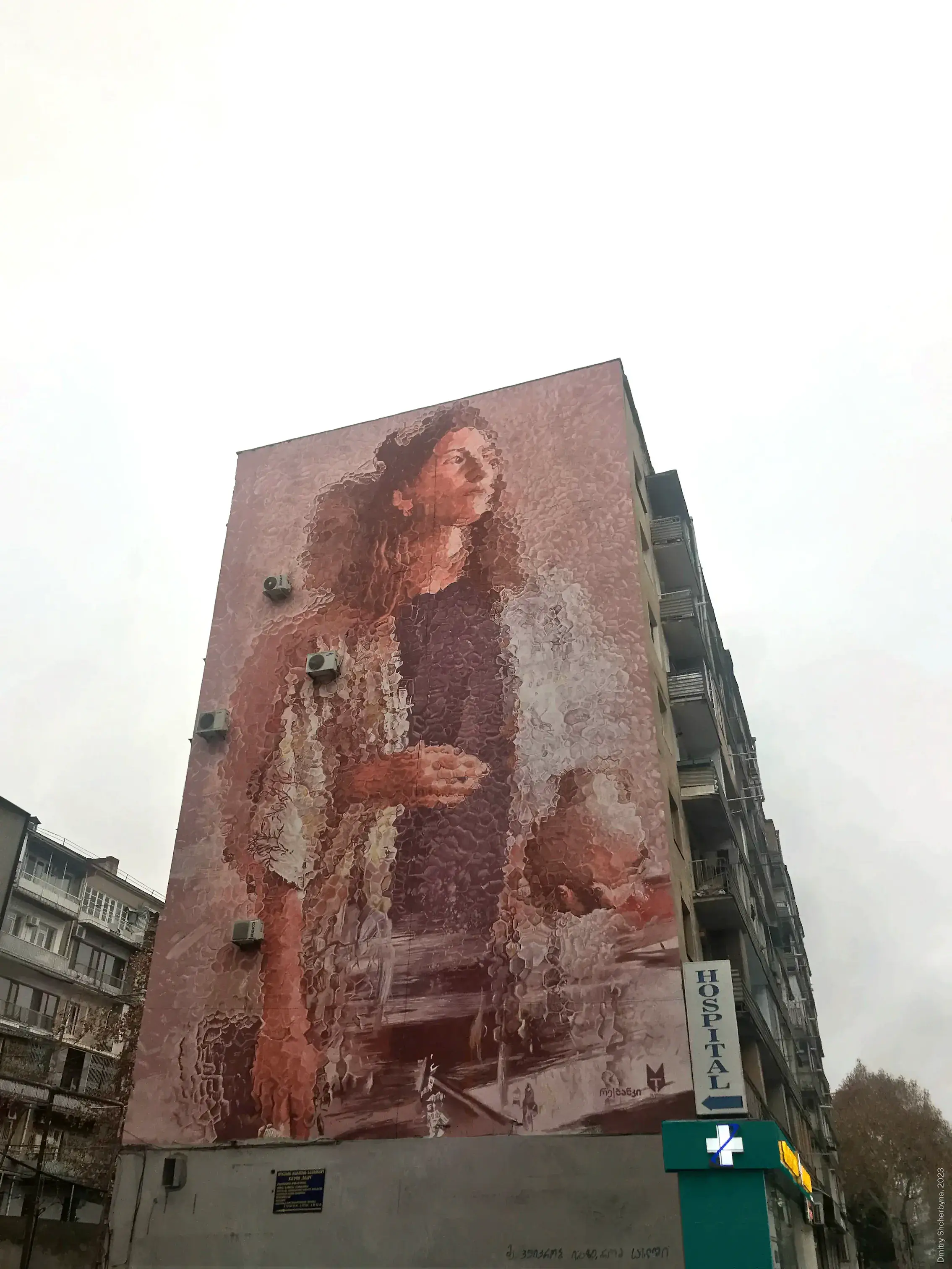 A full-building mural of a woman with long, curly hair rendered in a textured monochromatic palette of pink and mauve, looking upward with her hands gently clasped.