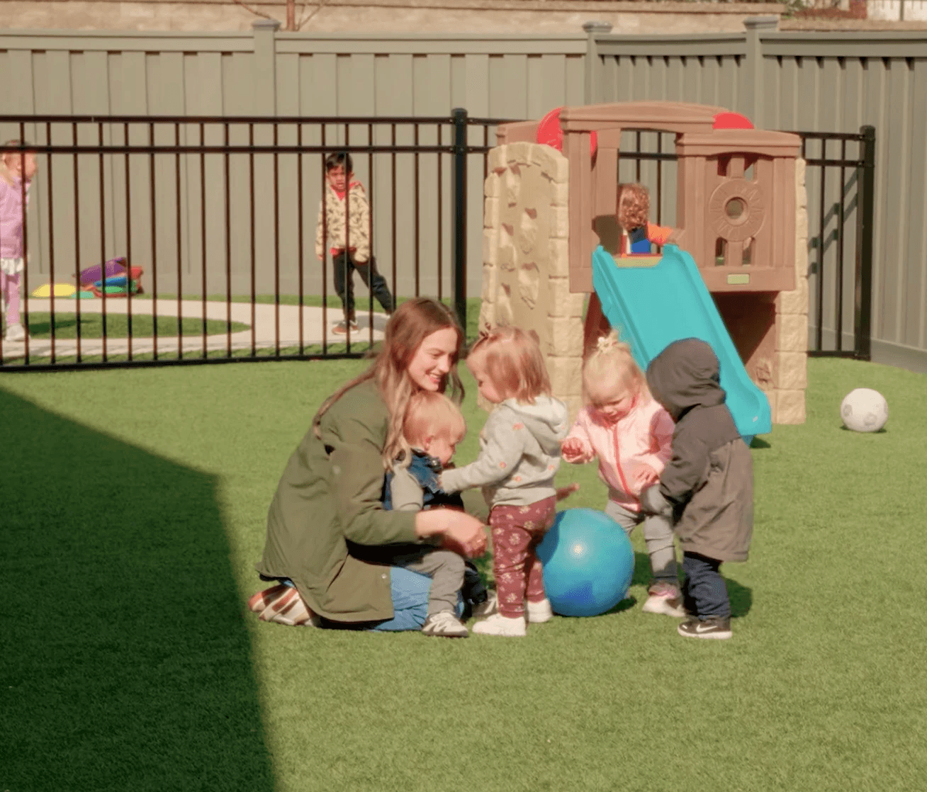 A woman plays with four children and a ball at a fenced in playground.