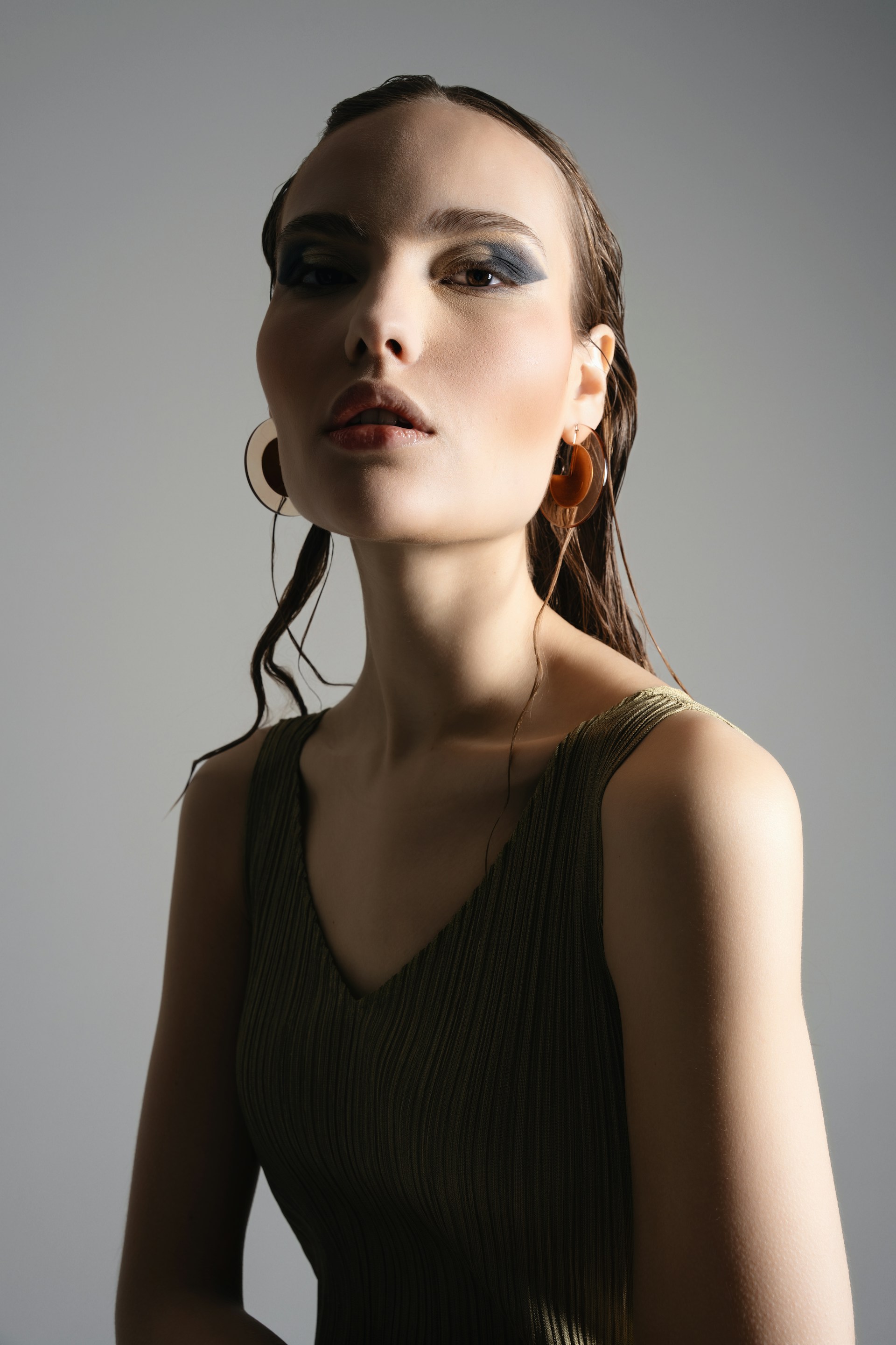 A low-angle portrait of the woman featuring large, translucent brown circular earrings. She has bold eye makeup and wet-look hair, posed against a plain grey background with sharp shadows.