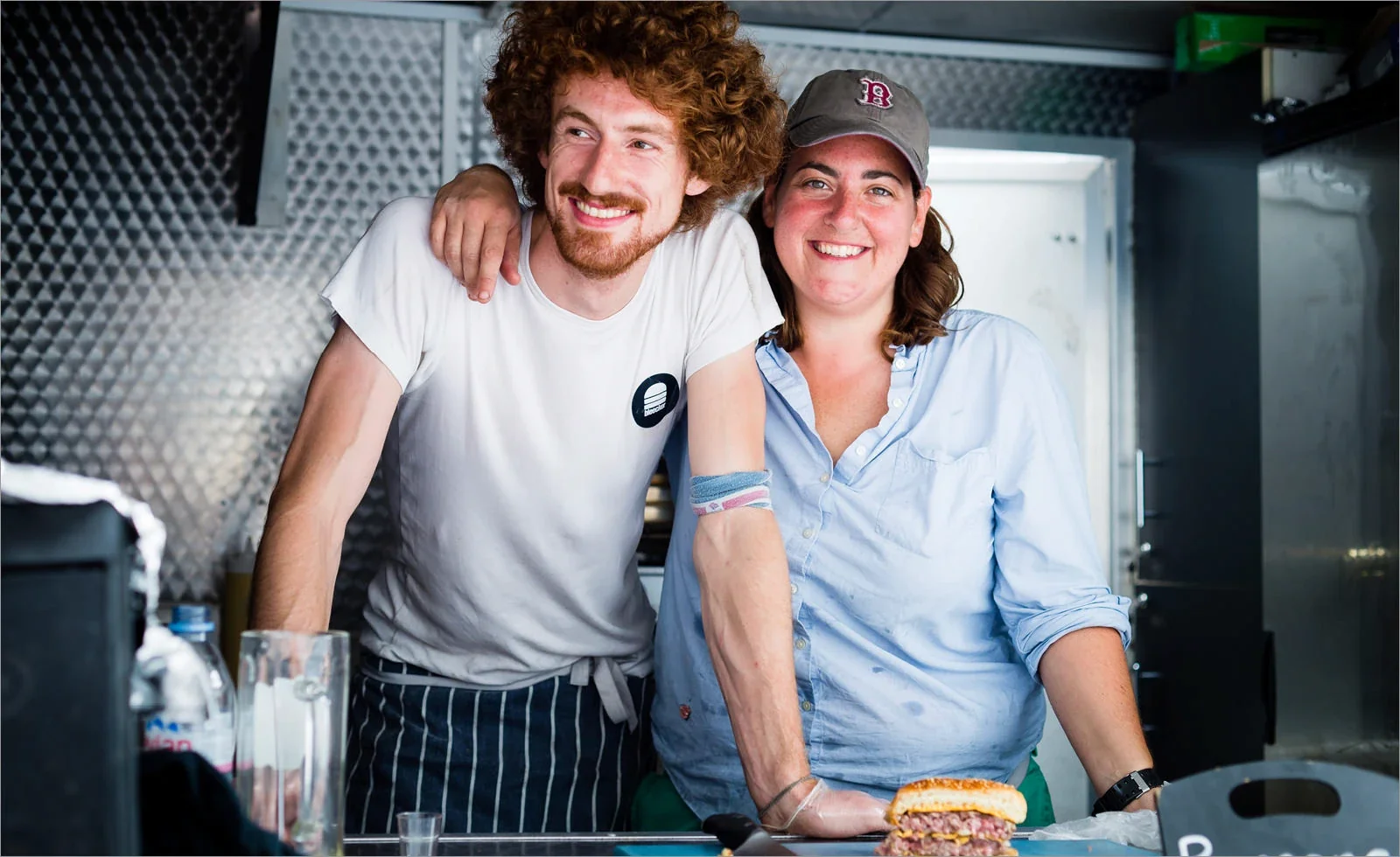 Two smiling food service workers stand behind a counter with a burger, wearing casual attire in a modern kitchen setting.