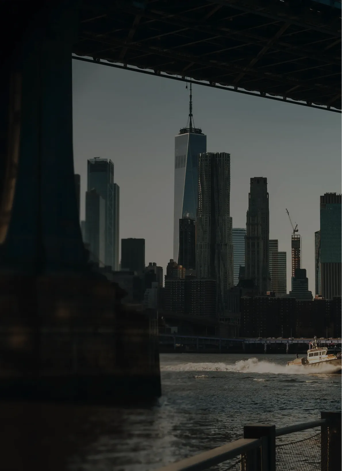 View of the Lower Manhattan skyline and One World Trade Center from beneath the Brooklyn Bridge, with a boat crossing the East River.