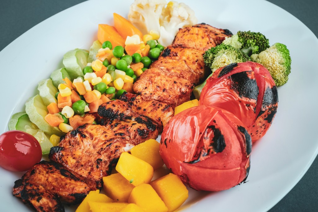 A delicious plate of grilled chicken skewers paired with a vibrant assortment of vegetables including broccoli, peas, carrots, tomatoes, cauliflower, and corn, showcasing a healthy and colorful meal option.