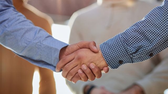 Aperto de mãos masculinas em camisas de manga longa; pessoas desfocadas ao fundo