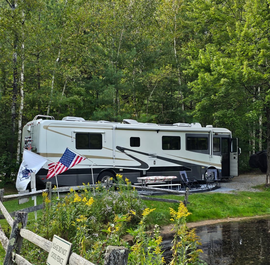 Large motorhome parked at a wooded waterfront site with flags and a picnic table