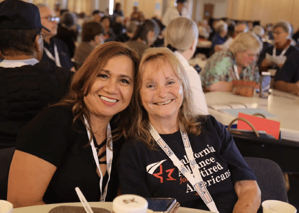 Two women are smiling and sitting close together at a crowded indoor event. Both are wearing lanyards, and the woman on the right is wearing a navy blue shirt that reads “California Alliance for Retired Americans.” The background shows other attendees seated at round tables, engaged in conversation.