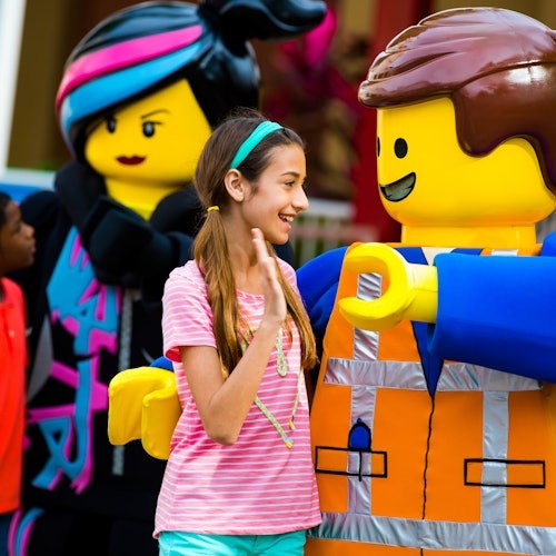 A girl in a pink shirt interacts happily with two life-sized Lego characters, one in construction attire and the other with colorful hair.