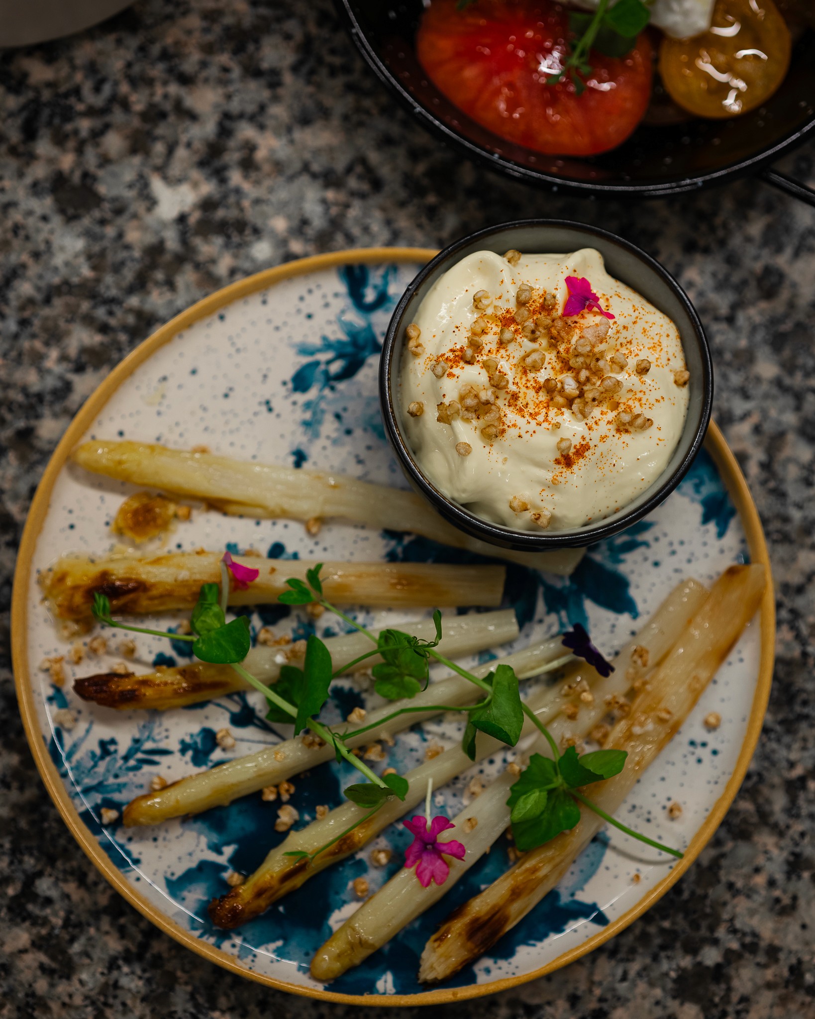 A dark green knitted scarf, a bouquet of orange gerbera daisies, and a newspaper lie on a granite ledge next to a body of water.