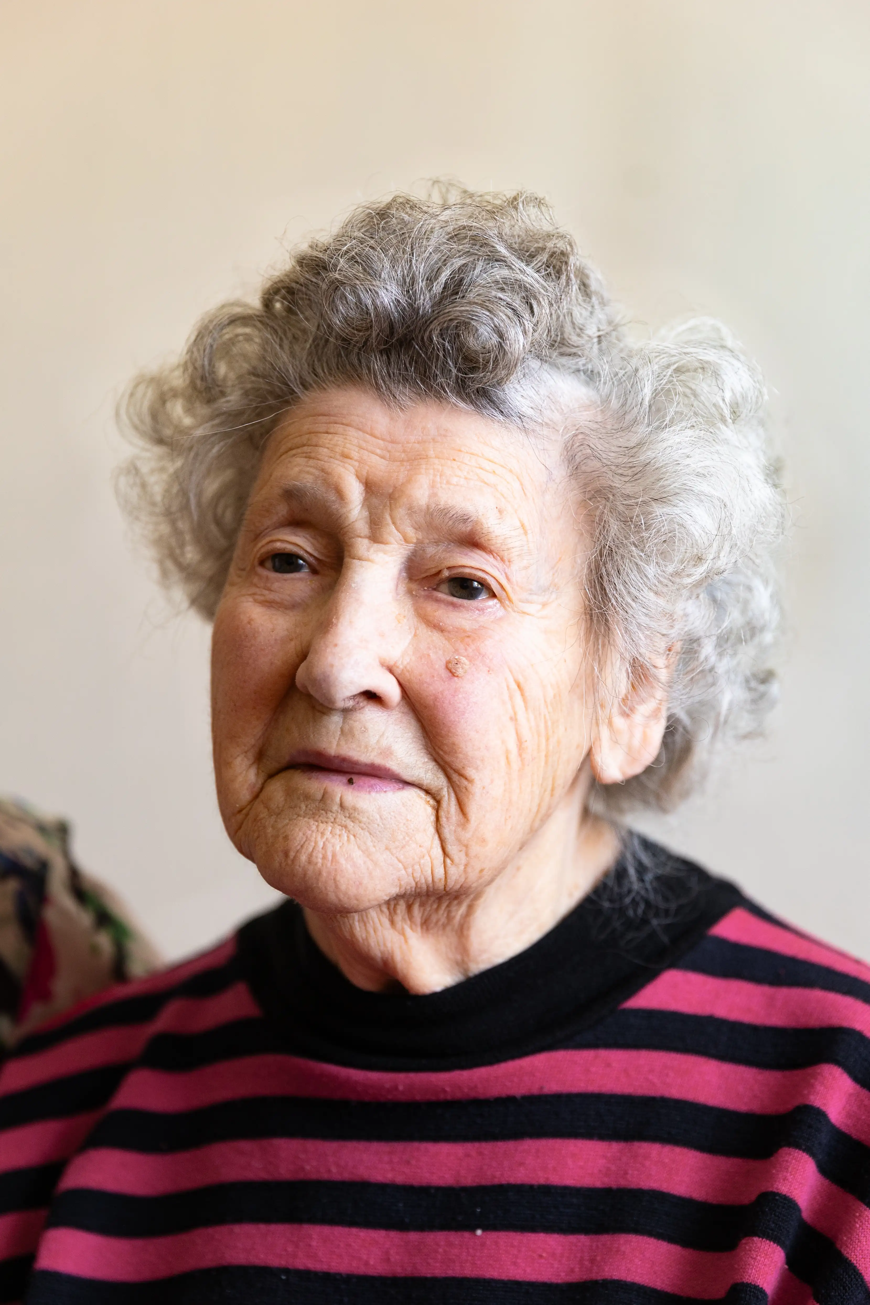 Starsza kobieta o siwych, kręconych włosach ze smutną miną. Ma na sobie bluzkę w czarno-różowe paski.