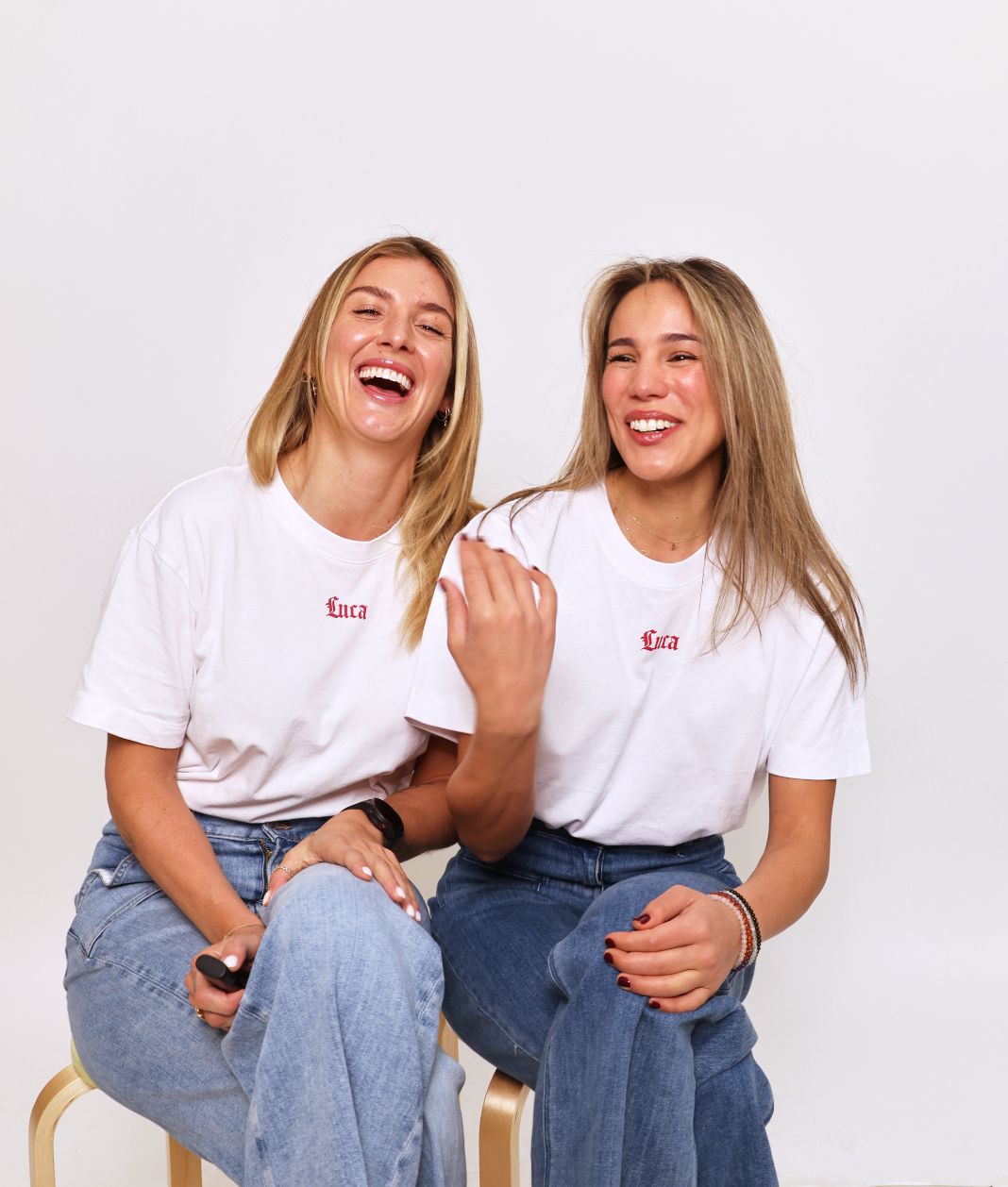 Two-smiling-women-sitting-together-and-laughing-in-a-studio- shot-They-are-both-wearing- matching-white-crewneck-t-shirts- that-feature-the-word-"Luca"-printed-in-small-red-script-on-the- chest-paired-with-denim-jeans