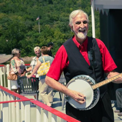 一位白发白胡须的男子在阳光明媚的户外露台上弹奏班卓琴，背景中有人在交谈。
