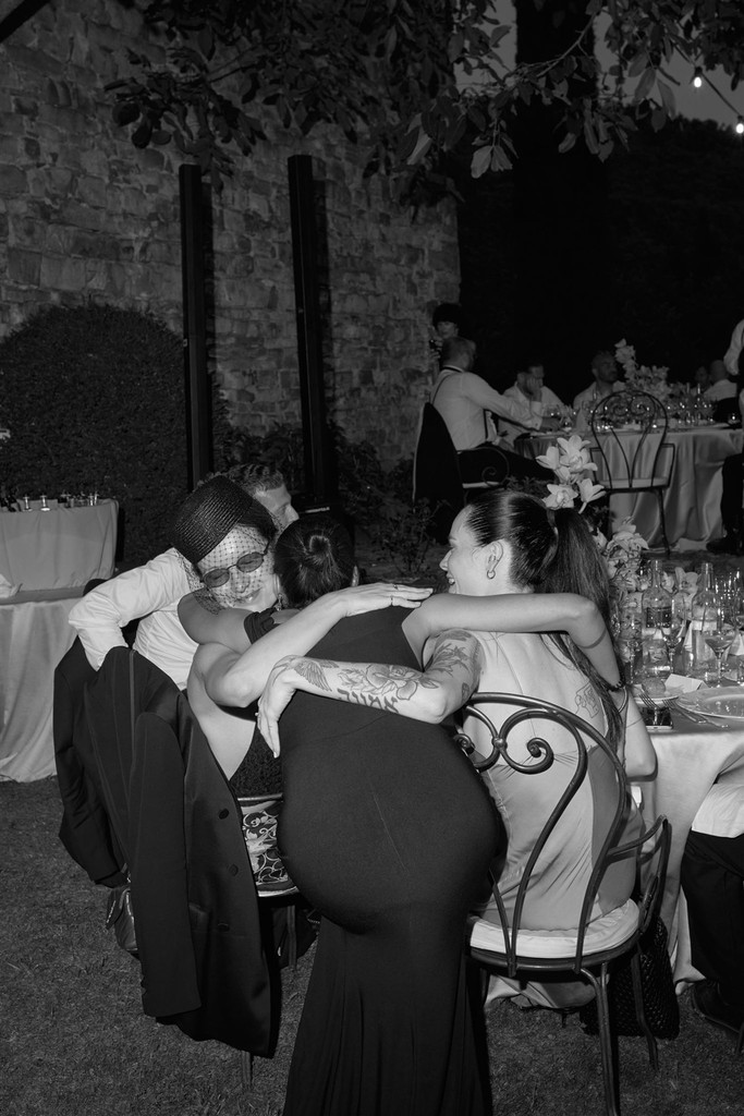Black-and-white flash portrait of guests hugging at the reception while sitting at their table