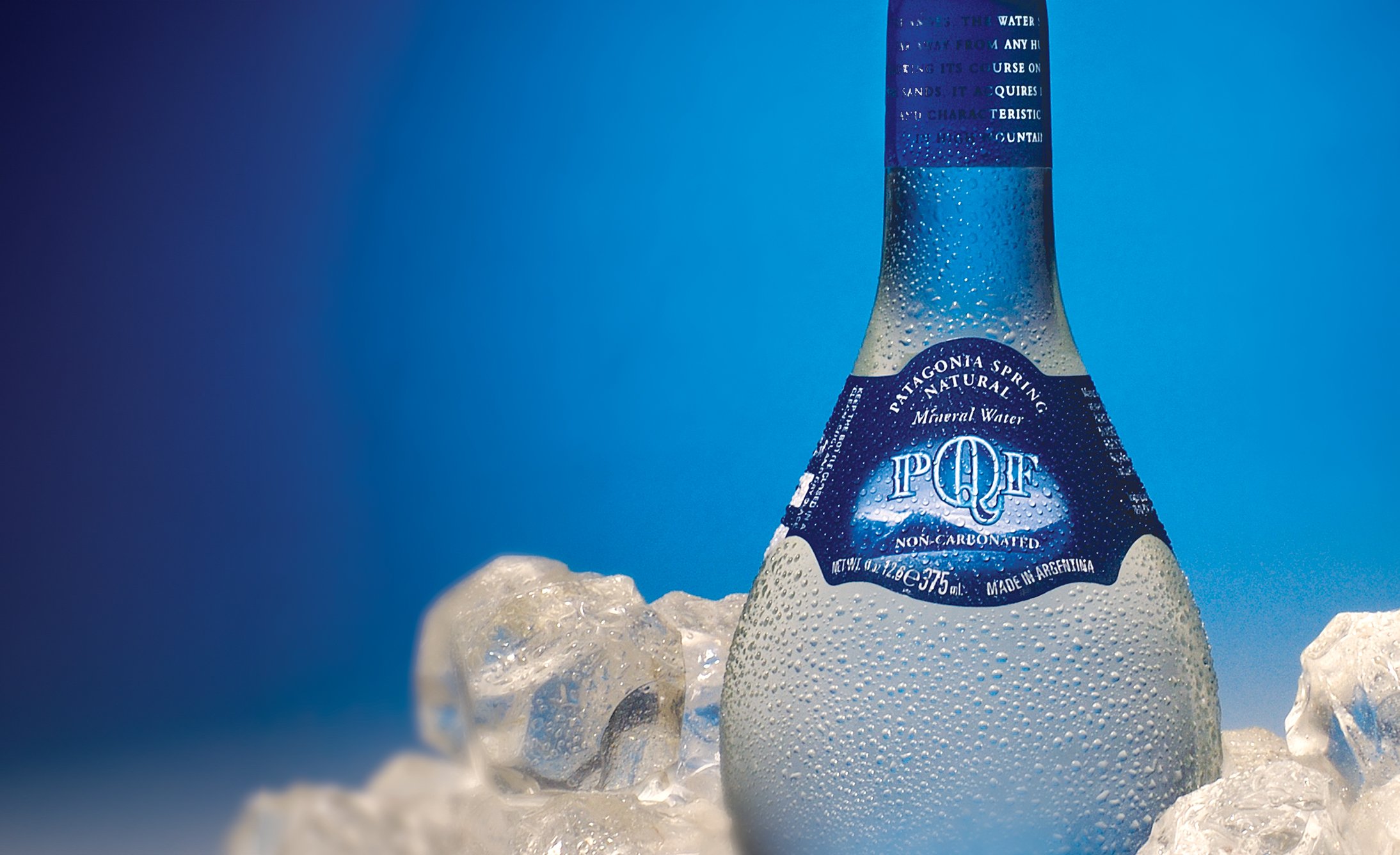 A blue-tinted bottle of mineral water labeled "Patagonia Spring" sits amid ice cubes on a blue background, conveying a refreshing and cool tone.