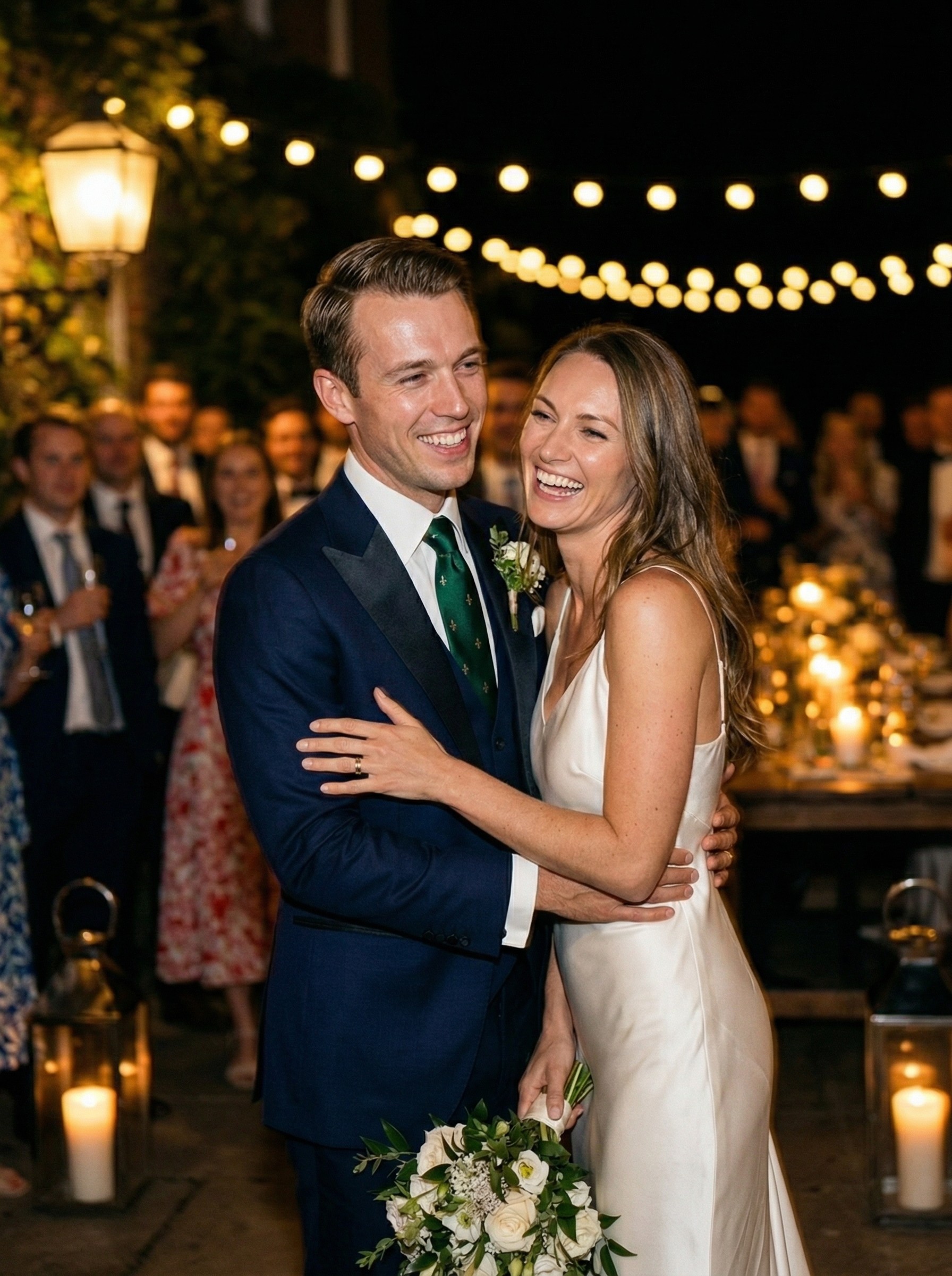 Bride and groom embracing and laughing during an outdoor evening wedding reception with string lights.