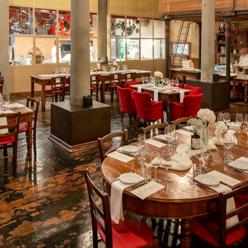 Isang restawrang may ilaw ng kandila, na may mga kahoy na mesang nakahanda para sa kainan, mga pulang upuan, at malalaking bintanang nakaharap sa kusina. Pinalalamutian ang mga mesa ng mga puting centerpiece na bulaklak.