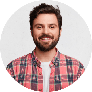 A man with a beard wearing a pink shirt, smiling and standing against a neutral background.