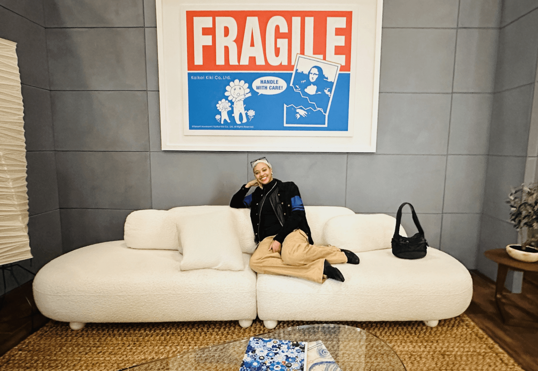 Jess smiling and lounging on a white couch beneath a large pop-art poster that reads “FRAGILE” in bold red and blue