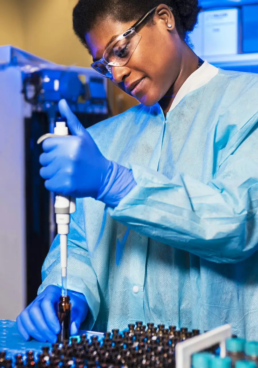 Scientist wearing safety goggles and blue lab attire, using a pipette to transfer liquid into a vial in a laboratory setting.