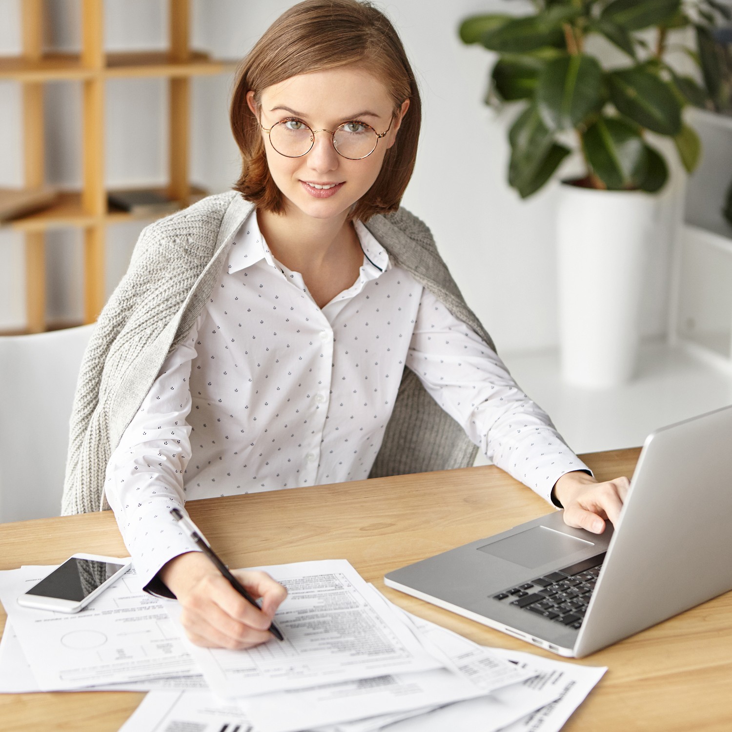 A person wearing glasses and a white polka-dotted shirt, draped with a gray sweater, sits at a wooden desk with a laptop, smartphone, and various documents, set against a modern office background with plants and shelving.