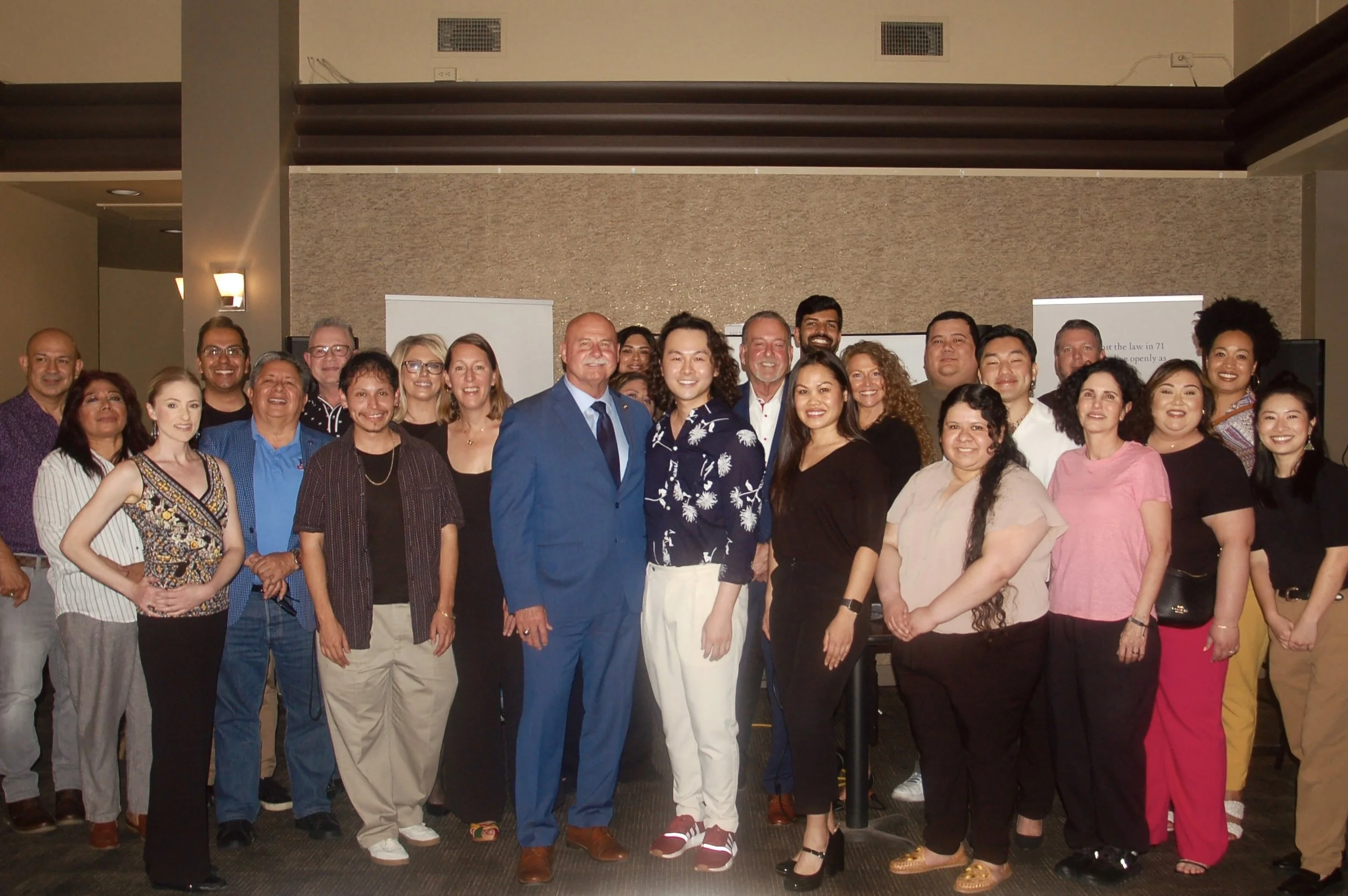 Um grupo diverso de pessoas, incluindo homens e mulheres de várias idades, posa junto em um ambiente interno durante um evento corporativo ou social, com dois banners verticais visíveis ao fundo.