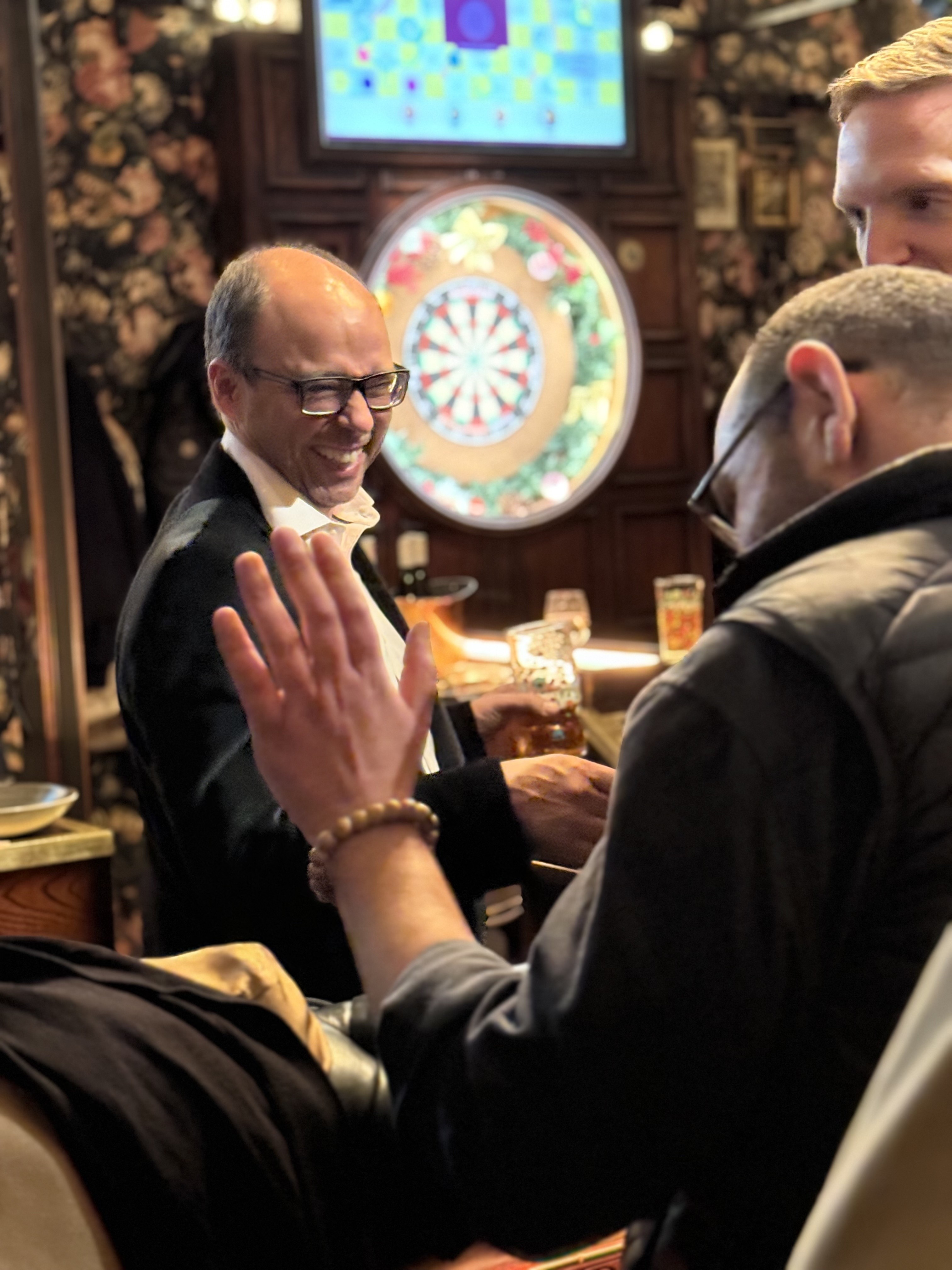 Two men are engaged in conversation in a cozy bar setting, with a decorative background and soft lighting.