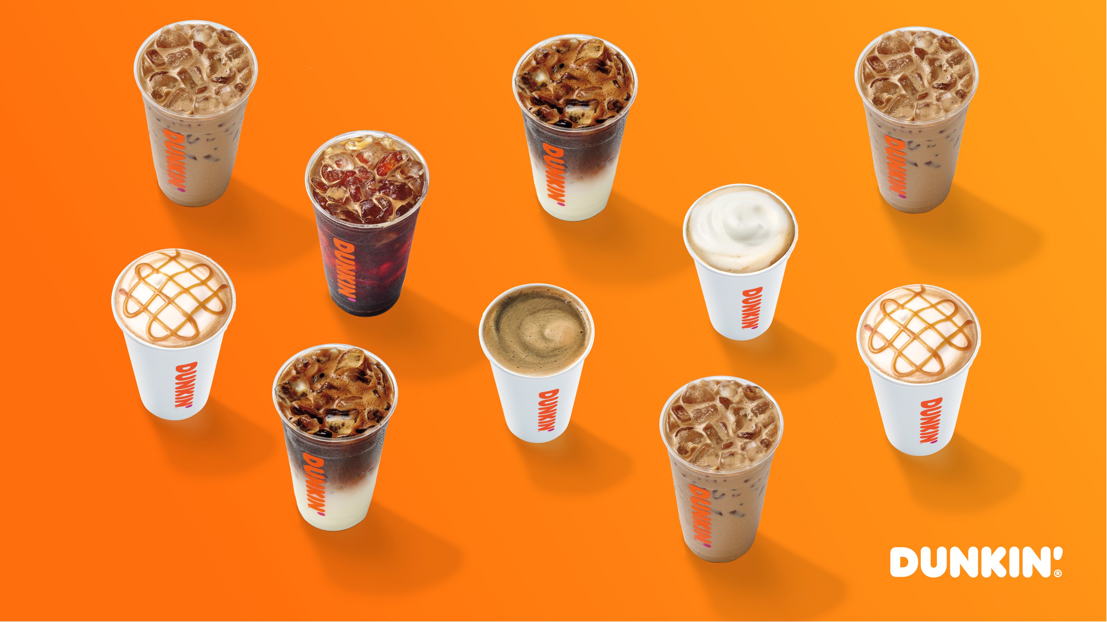 Selection of Dunkin’ hot and cold coffee drinks with frothy toppings and caramel drizzle designs, showcased on a vibrant orange surface.