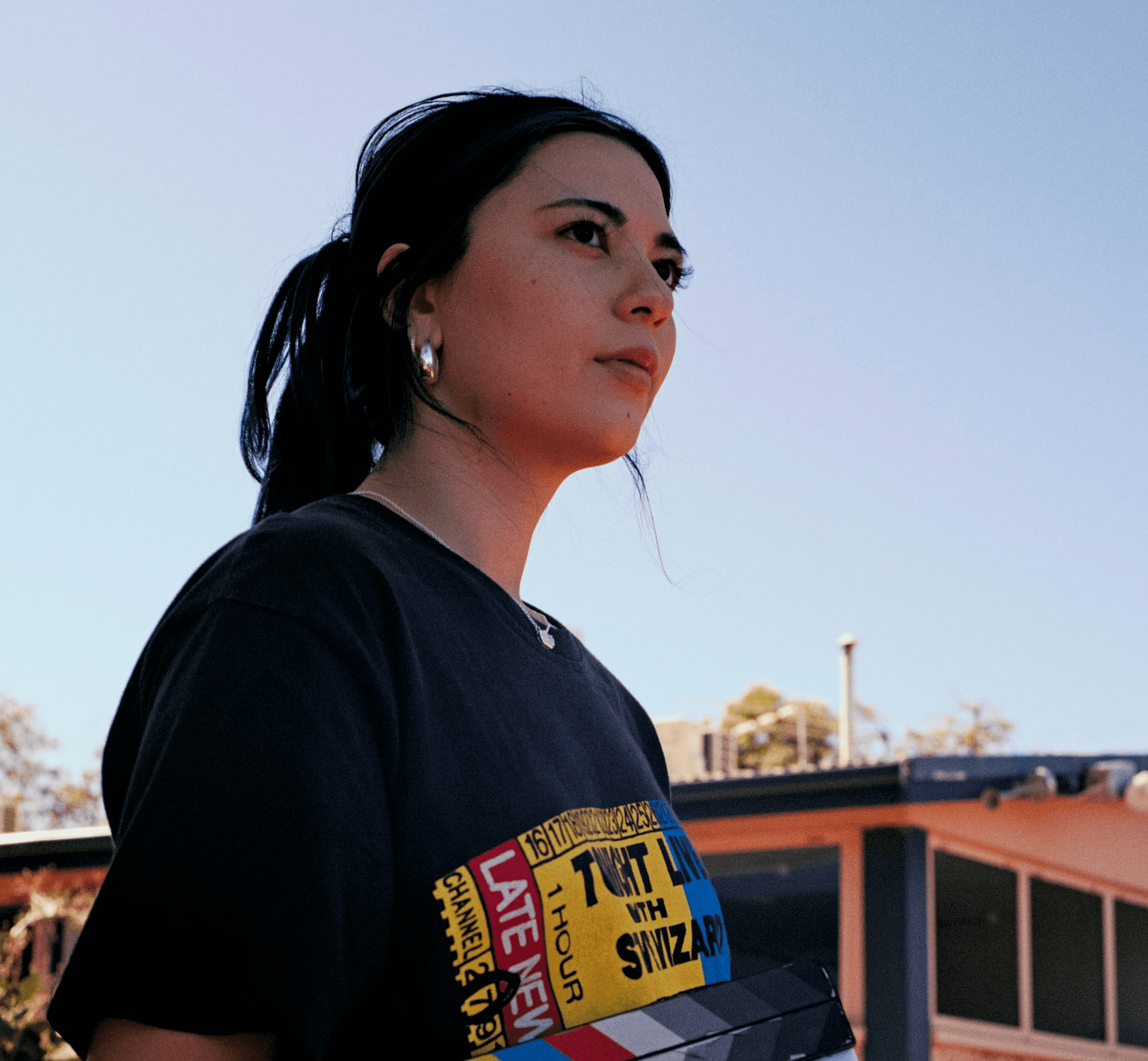 Young person with wavy, shoulder-length red hair, wearing a rust-colored vest over a beige shirt. A brown scarf is tied neatly at the neck. Neutral backdrop.