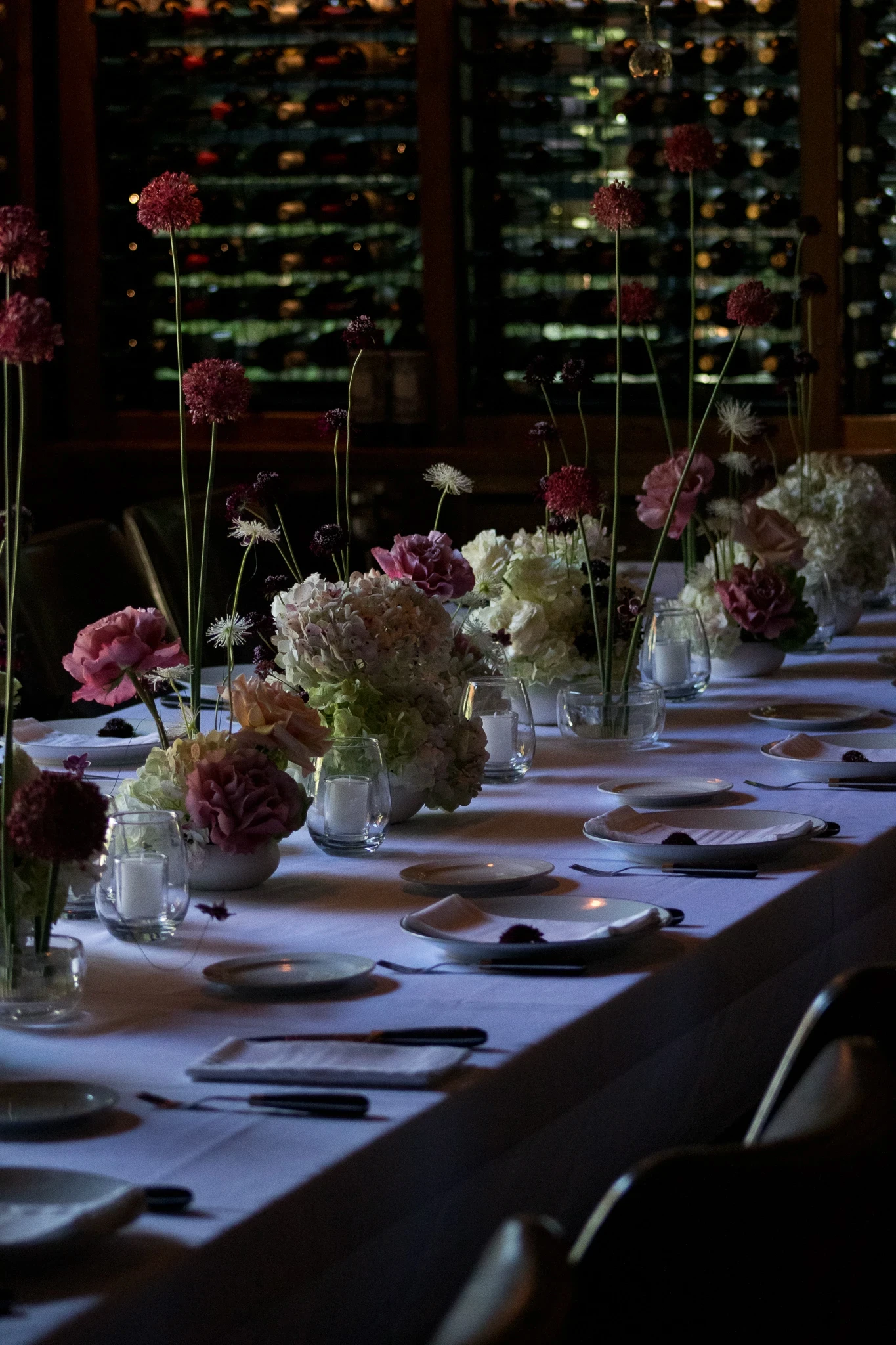 Fila de arreglos florales con aliums púrpura y rosas en mesa de recepción indoor con iluminación cálida e íntima.