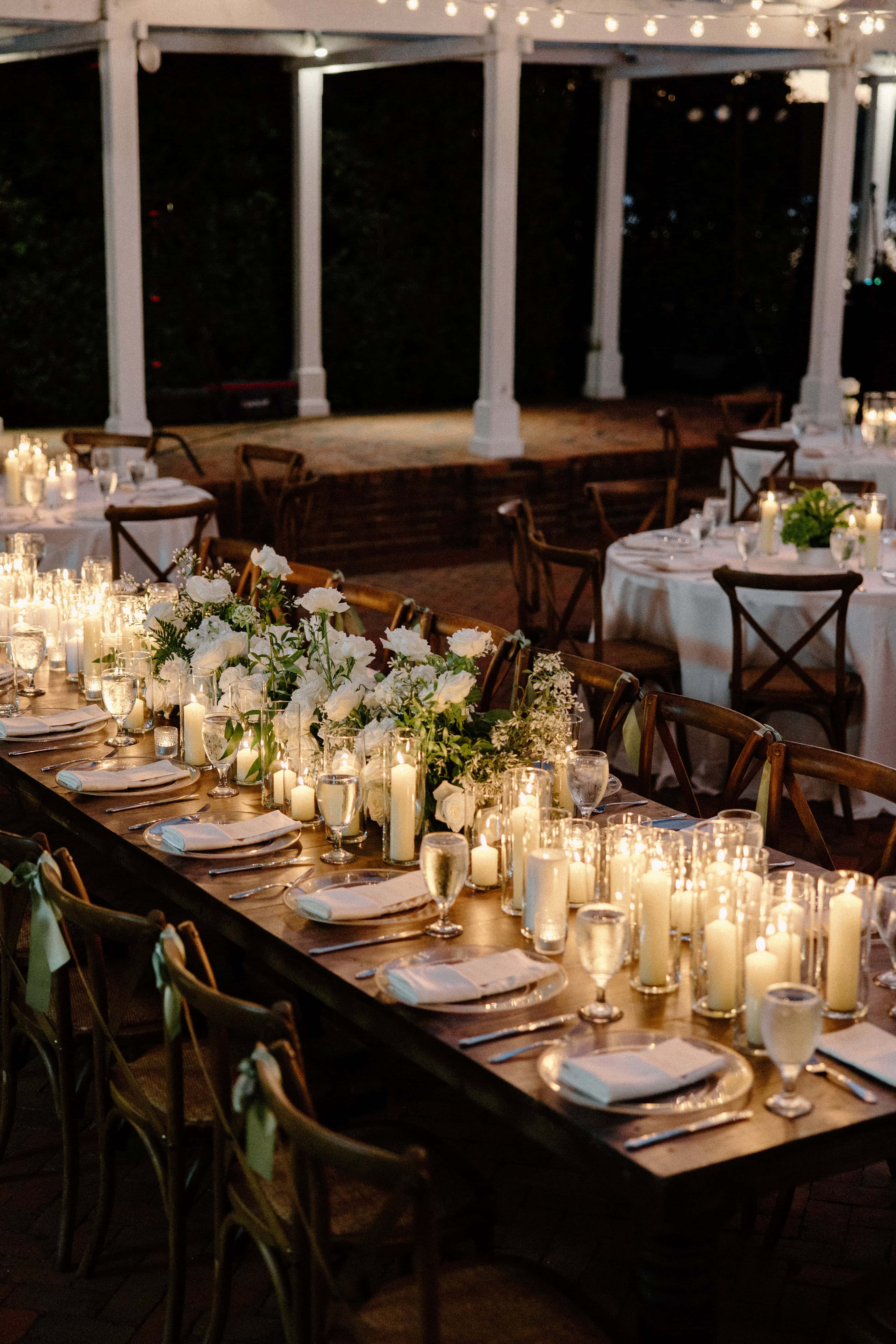 A nighttime shot of a long wedding reception table with floral centerpieces and many lit candles. In the background, there is a white pergola with string lights.