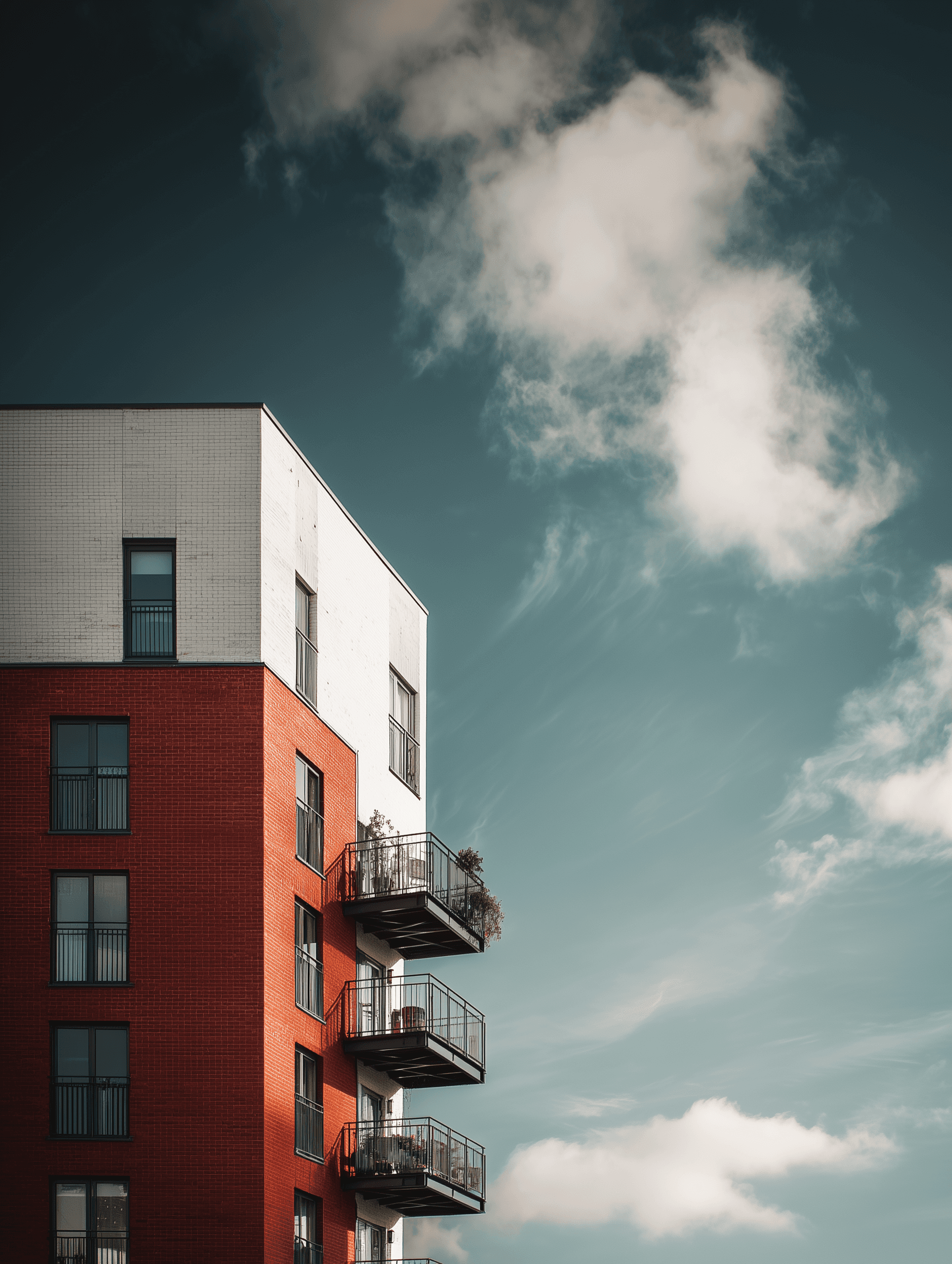 Photograph of a modern UK build-to-rent apartment building with a red brick lower facade and white upper facade. Several balconies extend outward with plants and outdoor furniture. The building is set against a dramatic sky with scattered clouds and soft sunlight.