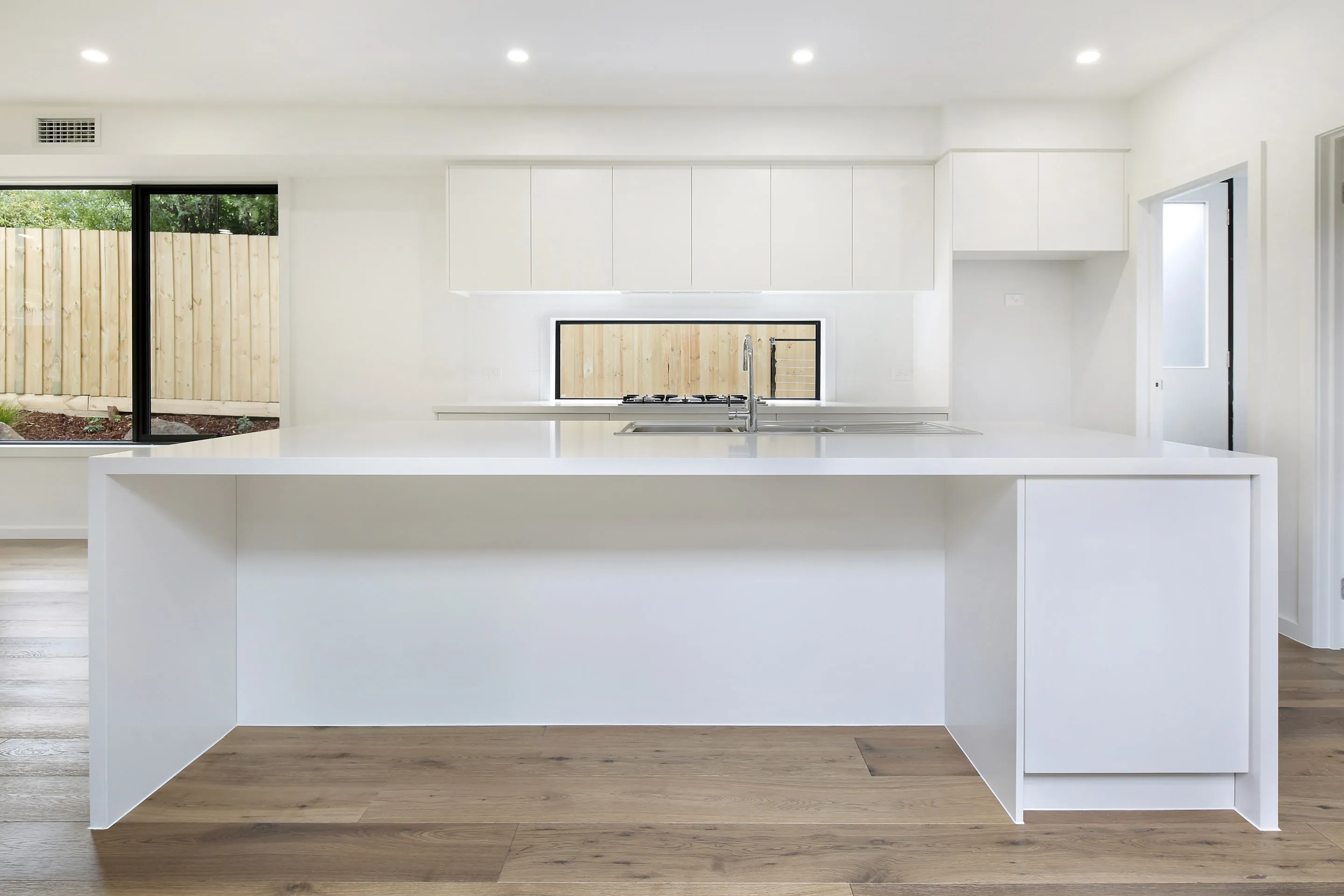 White kitchen with a long waterfall island, white overhead cabinetry, gas cooktop set under a window framing a timber fence, chrome tap and oak floors