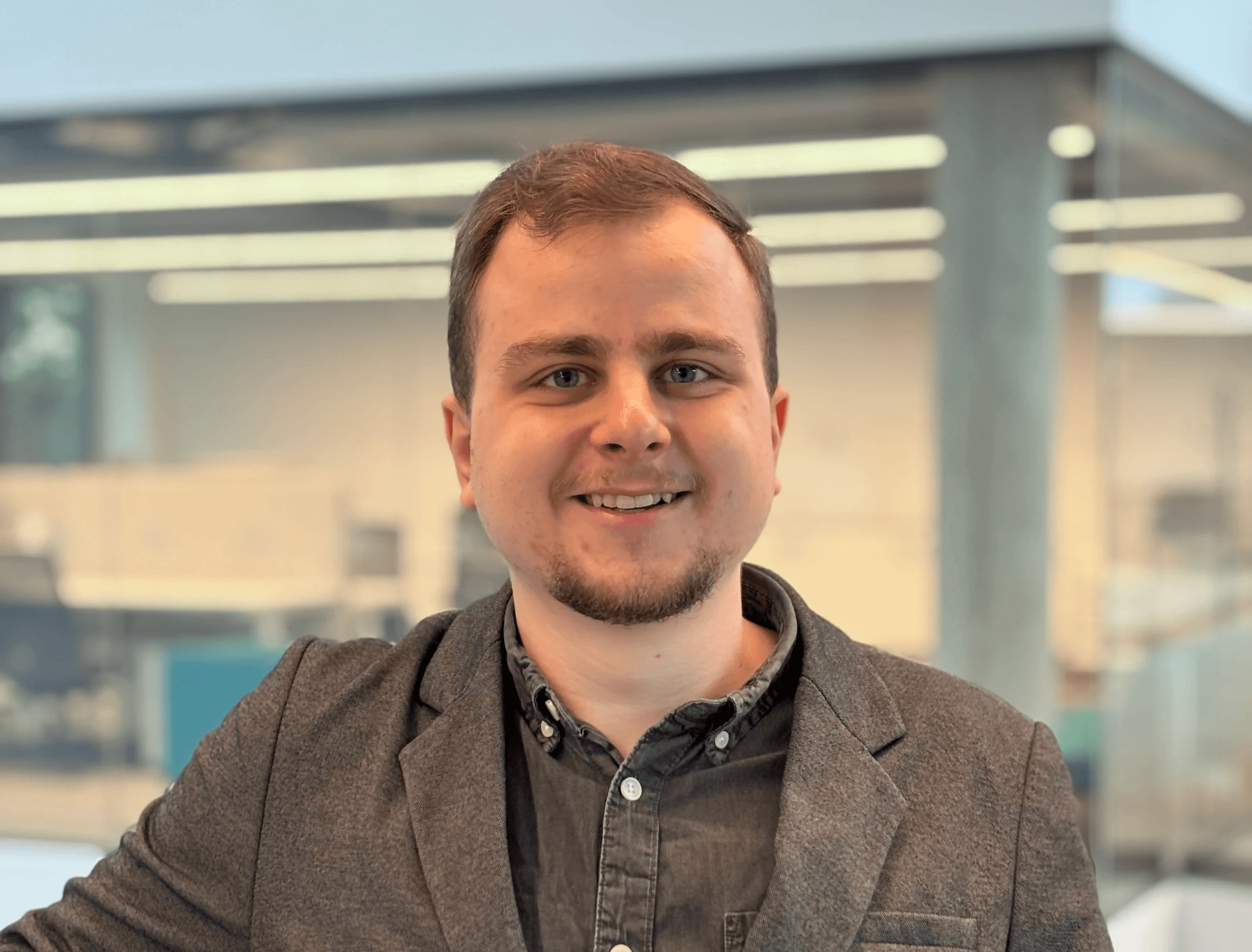 Smiling man with light brown hair and stubble, wearing a khaki shirt in natural light