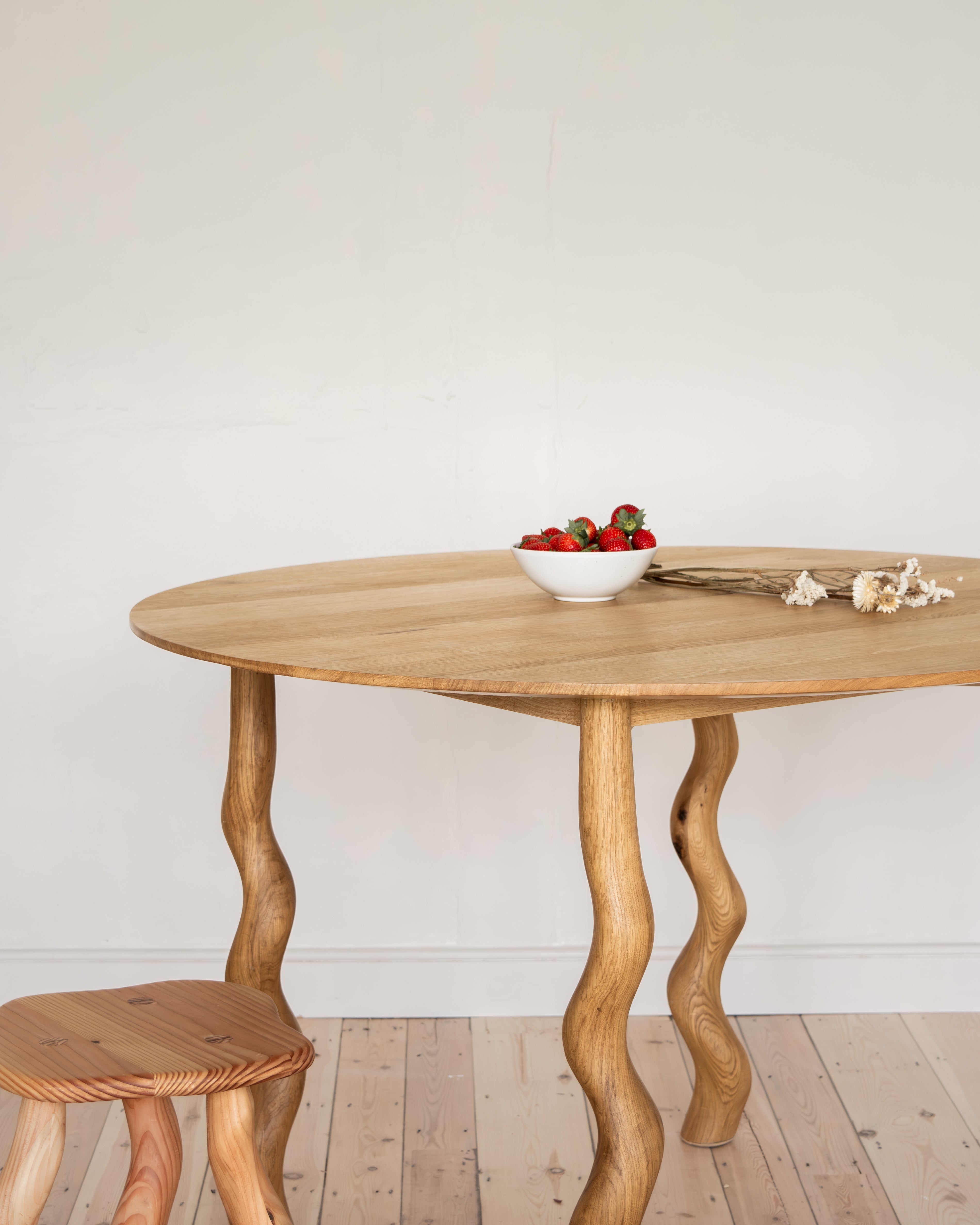 Wooden dining table with wavy legs on a wooden floor against a white wall.