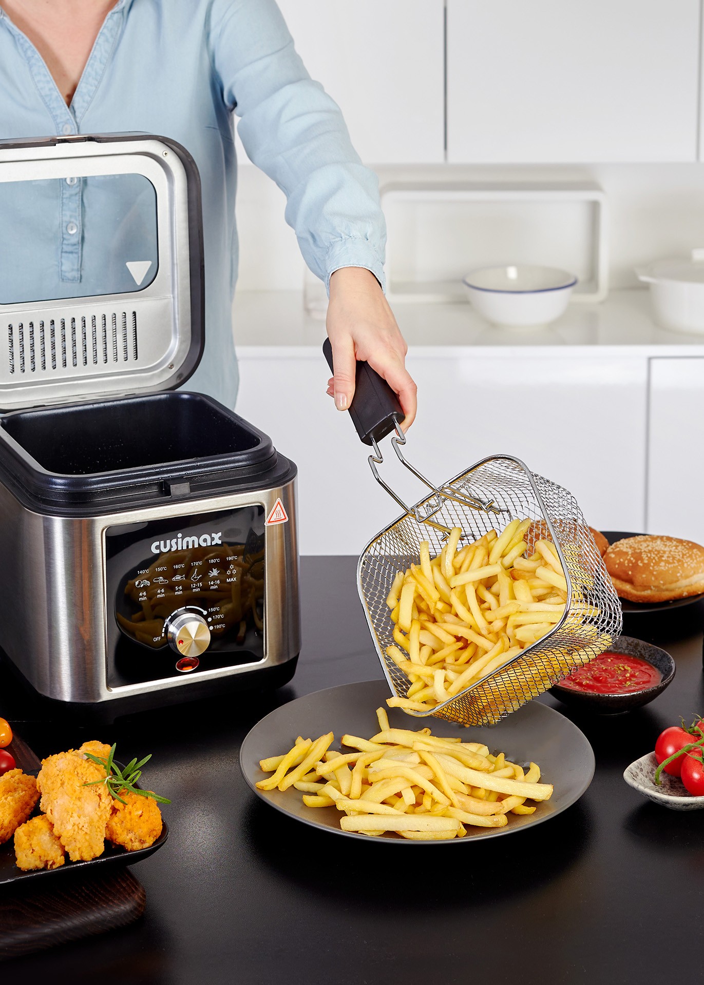 A person is using a stainless steel deep fryer to serve crispy French fries onto a plate, surrounded by fried chicken, tomatoes, and a hamburger bun on a kitchen counter.