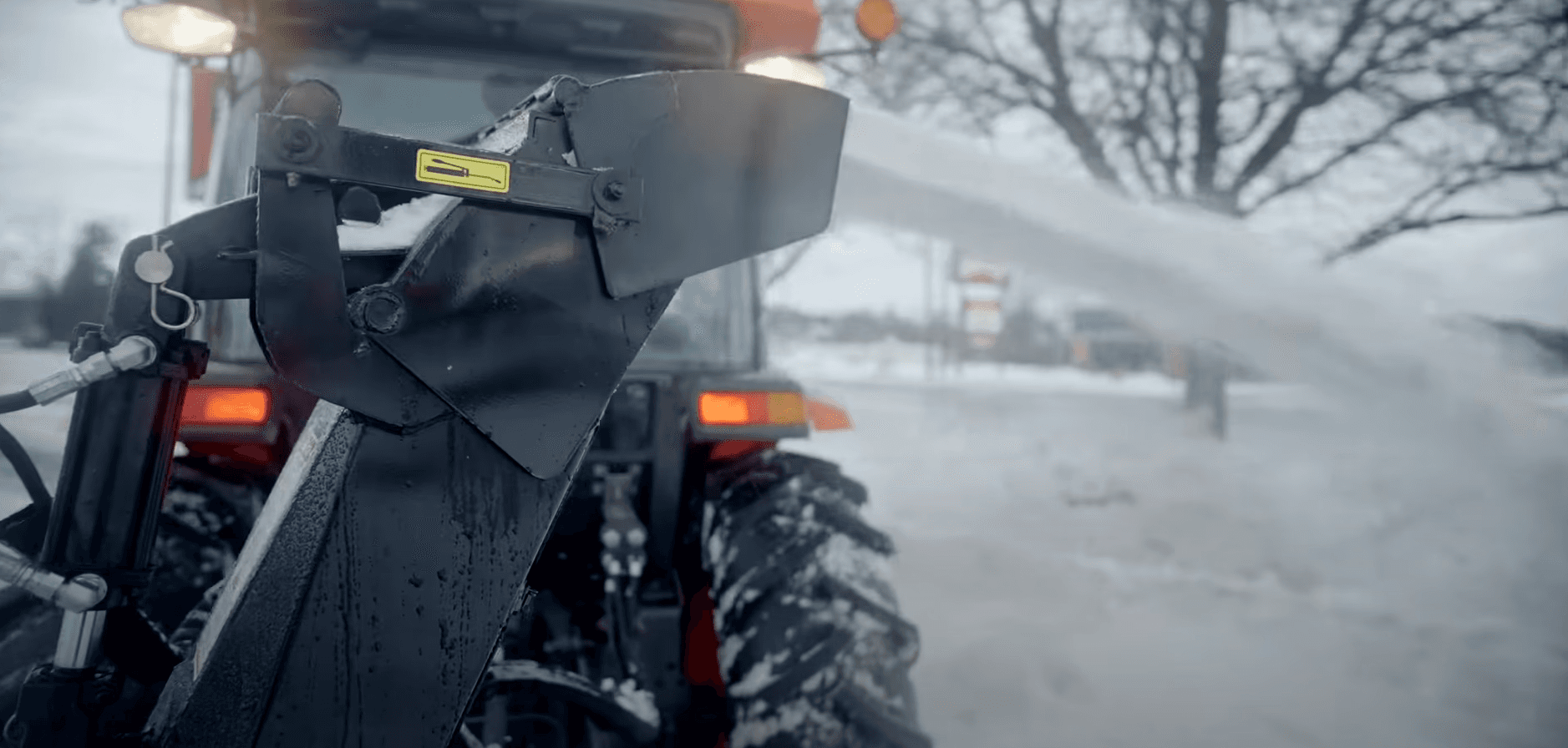Une souffleuse à neige attachée à un tracteur nettoie et éjecte activement la neige dans un environnement extérieur enneigé.