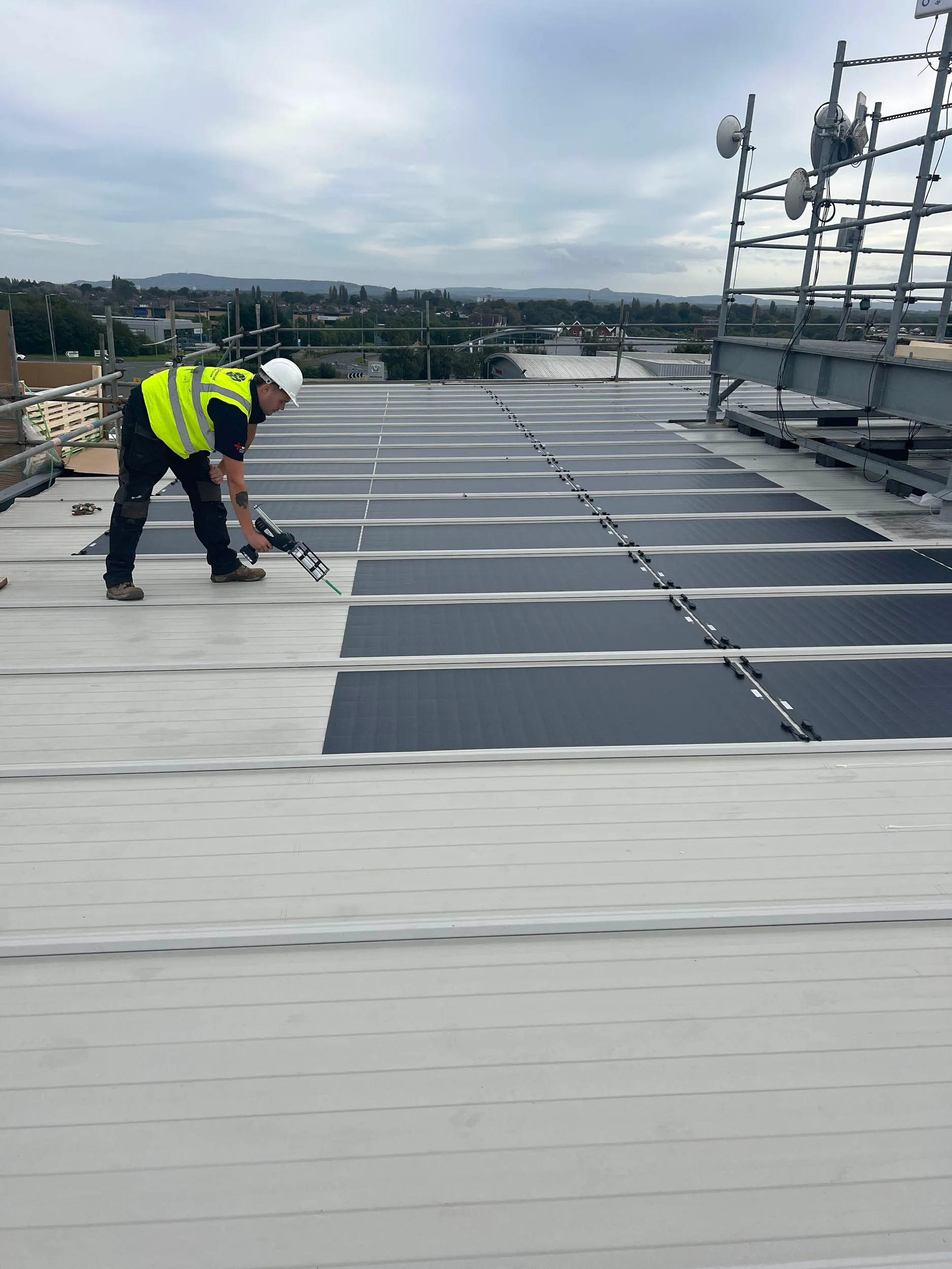 A worker in a high-visibility vest and hard hat installs solar panels on a large flat rooftop under a cloudy sky, with scaffolding visible in the background.