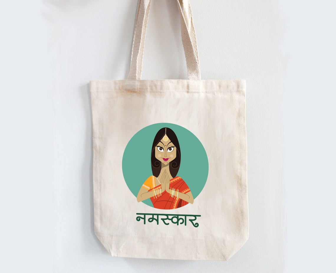 A cream-colored tote bag with off-white straps hangs against a white background. The bag features a circular graphic of a woman with dark hair, wearing a yellow-orange top and orange and red patterned fabric. She has her hands pressed together in a gesture resembling a prayer or greeting. Below the graphic, the word "नमस्कार" is printed in dark green text.