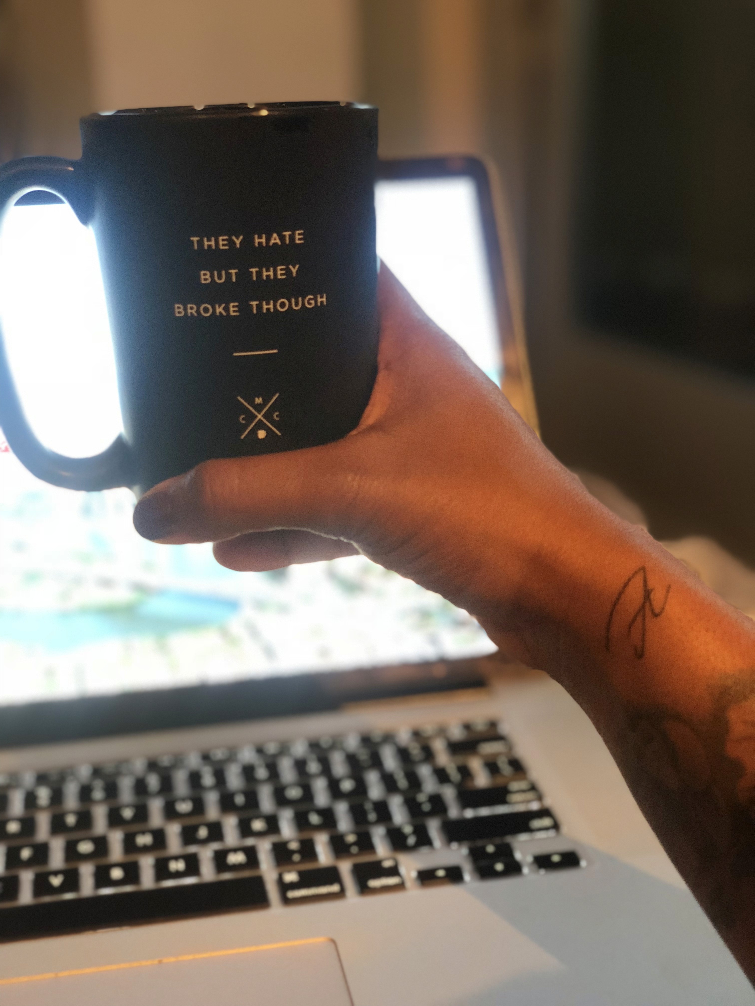 A hand holds a black mug in front of a laptop screen, with soft lighting in the background.