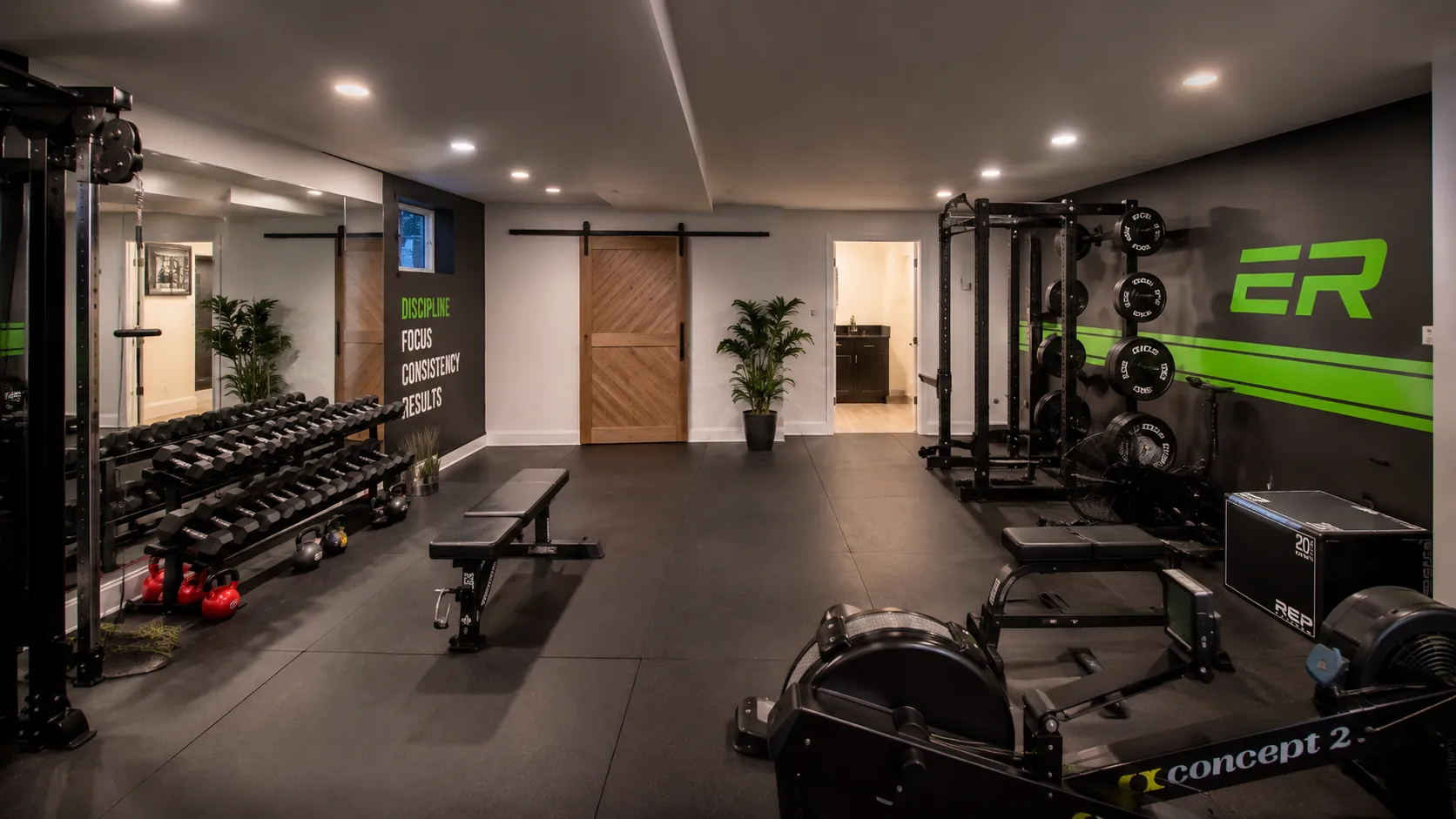Modern home gym basement with complete dumbbell set, Smith machine, squat rack, motivational green neon branding, and dark athletic flooring with bright LED ceiling lighting.