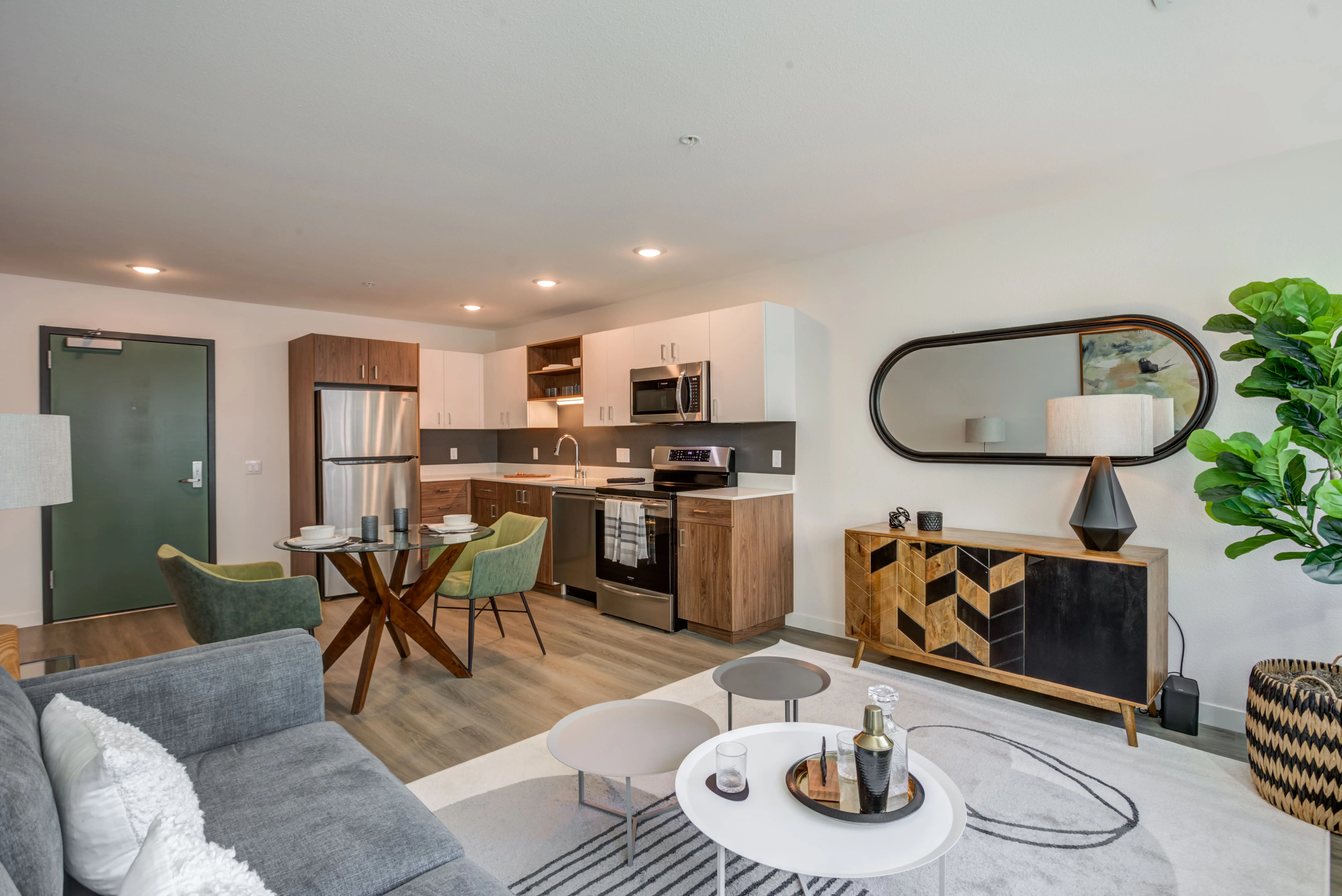 Modern open-concept apartment interior in Midtown Sacramento featuring a living area with a gray sofa, nested coffee tables, and a rug. The kitchen includes stainless steel appliances, white and wood cabinetry, and a small dining table with green chairs. Warm lighting sets a cozy ambiance.