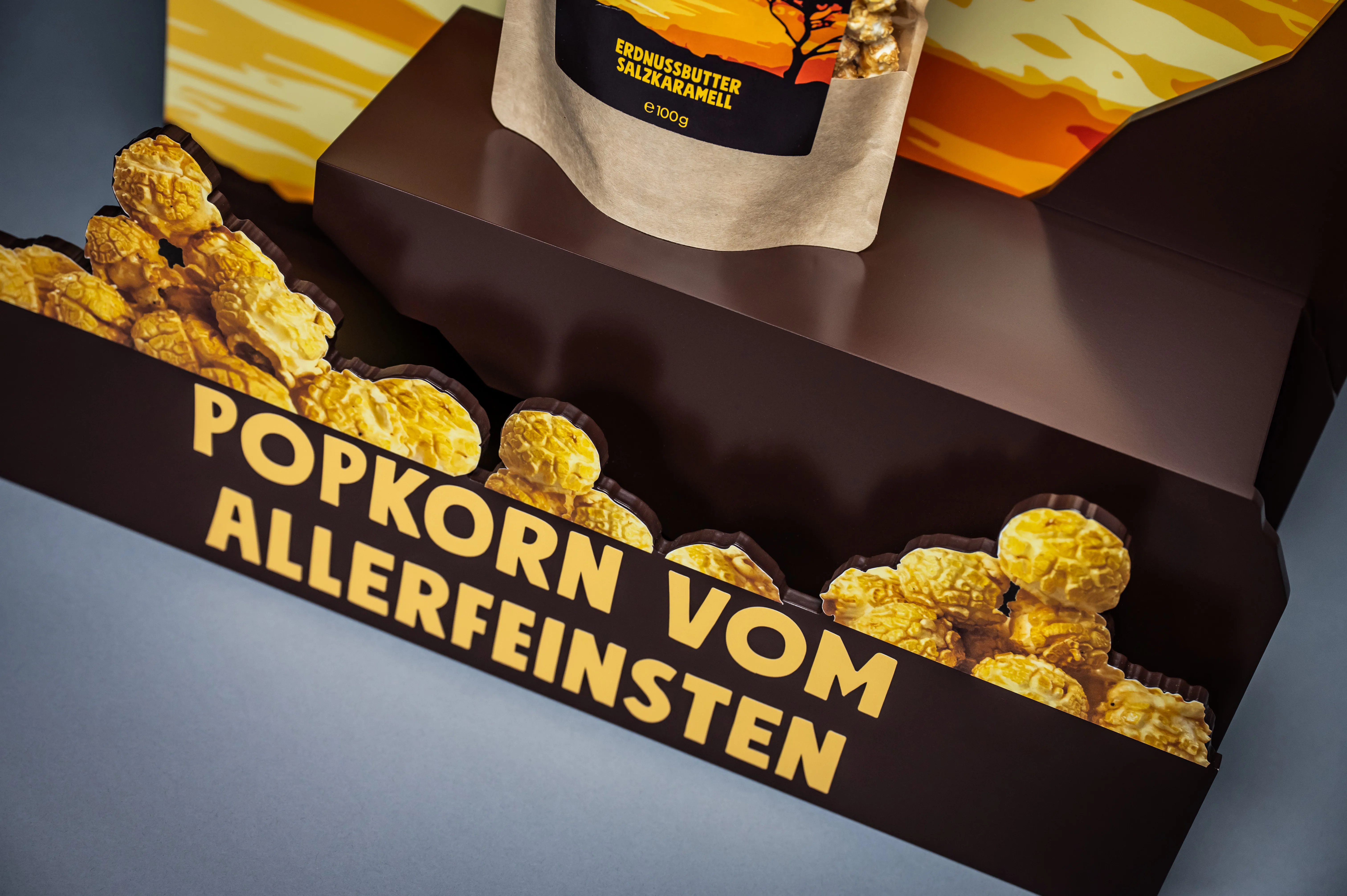 Detail of the KNALLE display tray with yellow Popkorn vom Allerfeinsten lettering on cut-out popcorn silhouettes and a peanut butter salt caramel bag above.