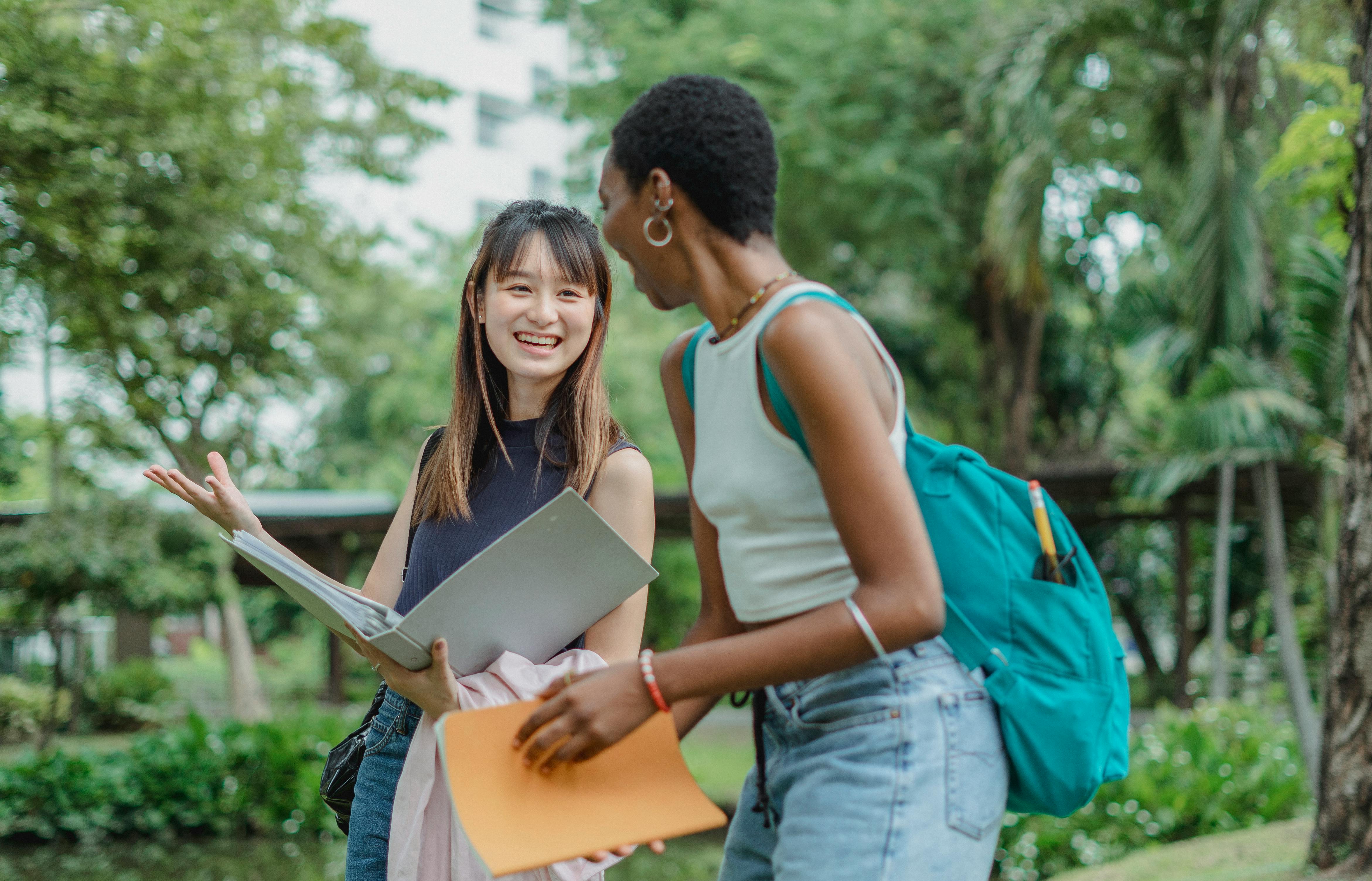 Three students with backpacks and folders chat and smile outside a brick building with a prominent glass entrance.