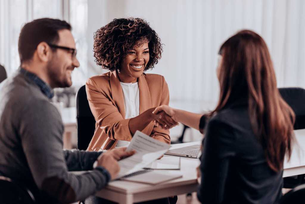 Três pessoas em uma mesa: à esquerda, um jovem de barba e cabelos loiros, sorrindo. Ao seu lado, uma mulher negra em blazer, com cabelos cacheados na altura das orelhas, sorrindo enquanto estende o braço sobre a mesa para apertar a mão de uma mulher à direita. Ambas estão sorrindo, transmitindo uma atmosfera de cordialidade e interação positiva.