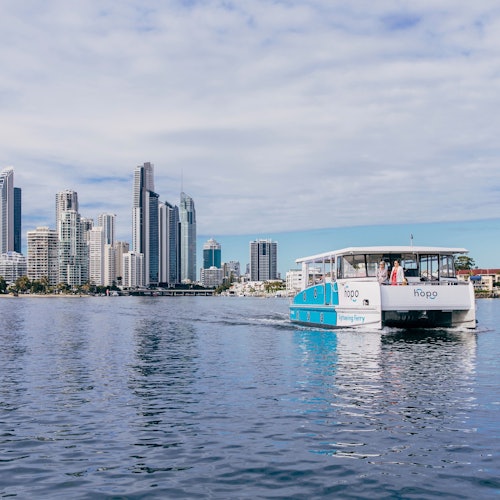 A ferry with people aboard moves across calm water with a city skyline of tall buildings and blue sky in the background.