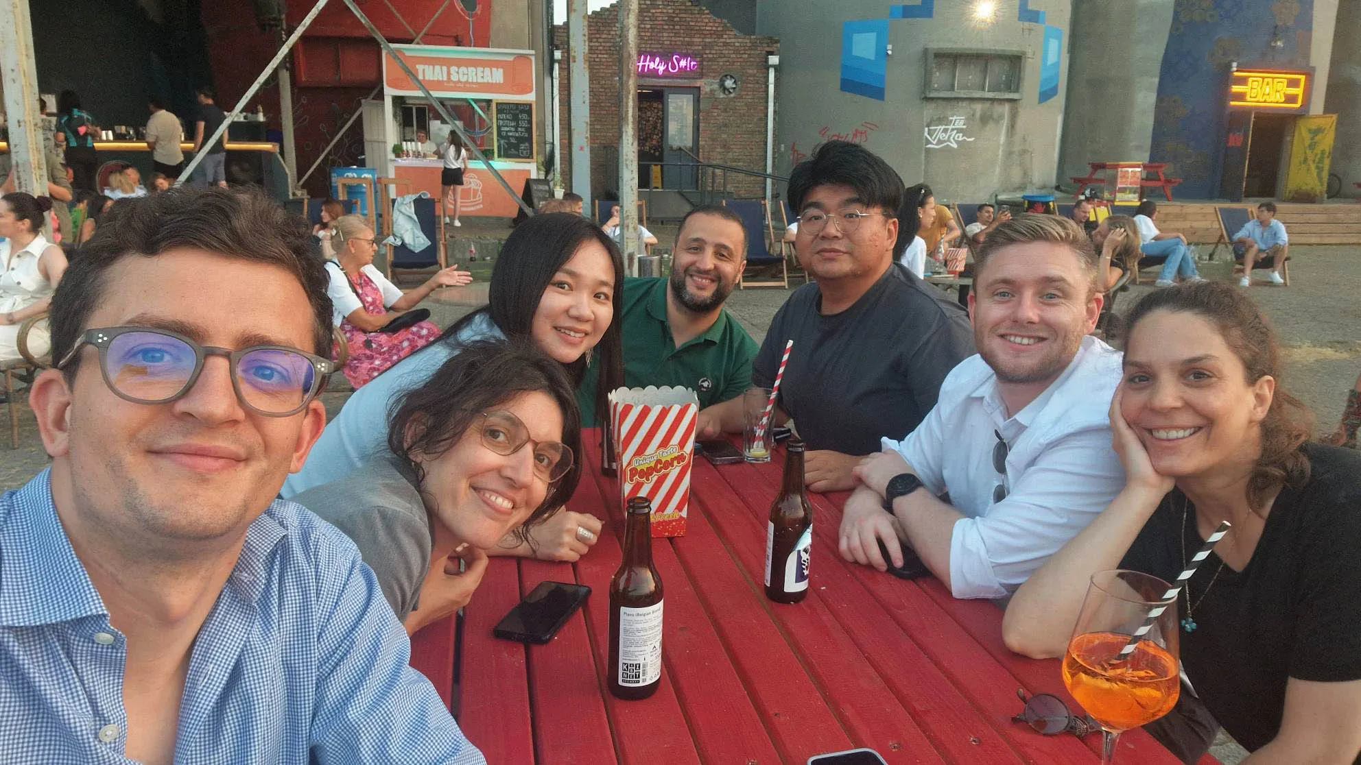 A group of seven people smiling around a table with drinks, enjoying time together outdoors.