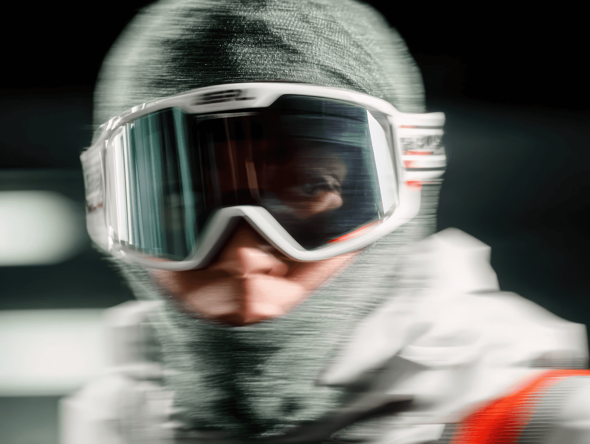 Close-up of a person in a green balaclava and white ski goggles, with a dynamic and slightly blurred effect, suggesting motion.