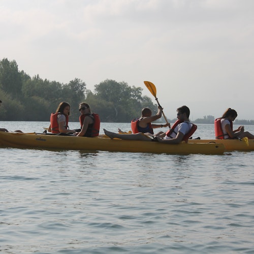 Cinco personas con chalecos salvavidas remando en un kayak amarillo en un lago tranquilo con una línea de árboles al fondo.