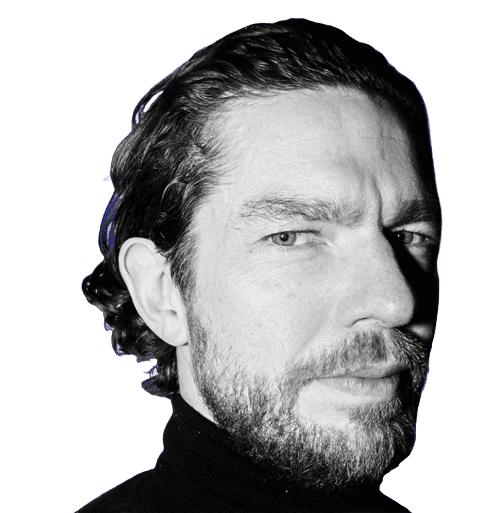 A young person with curly hair smiles at the camera while sitting in front of a laptop against a dark background. (Background Removed)