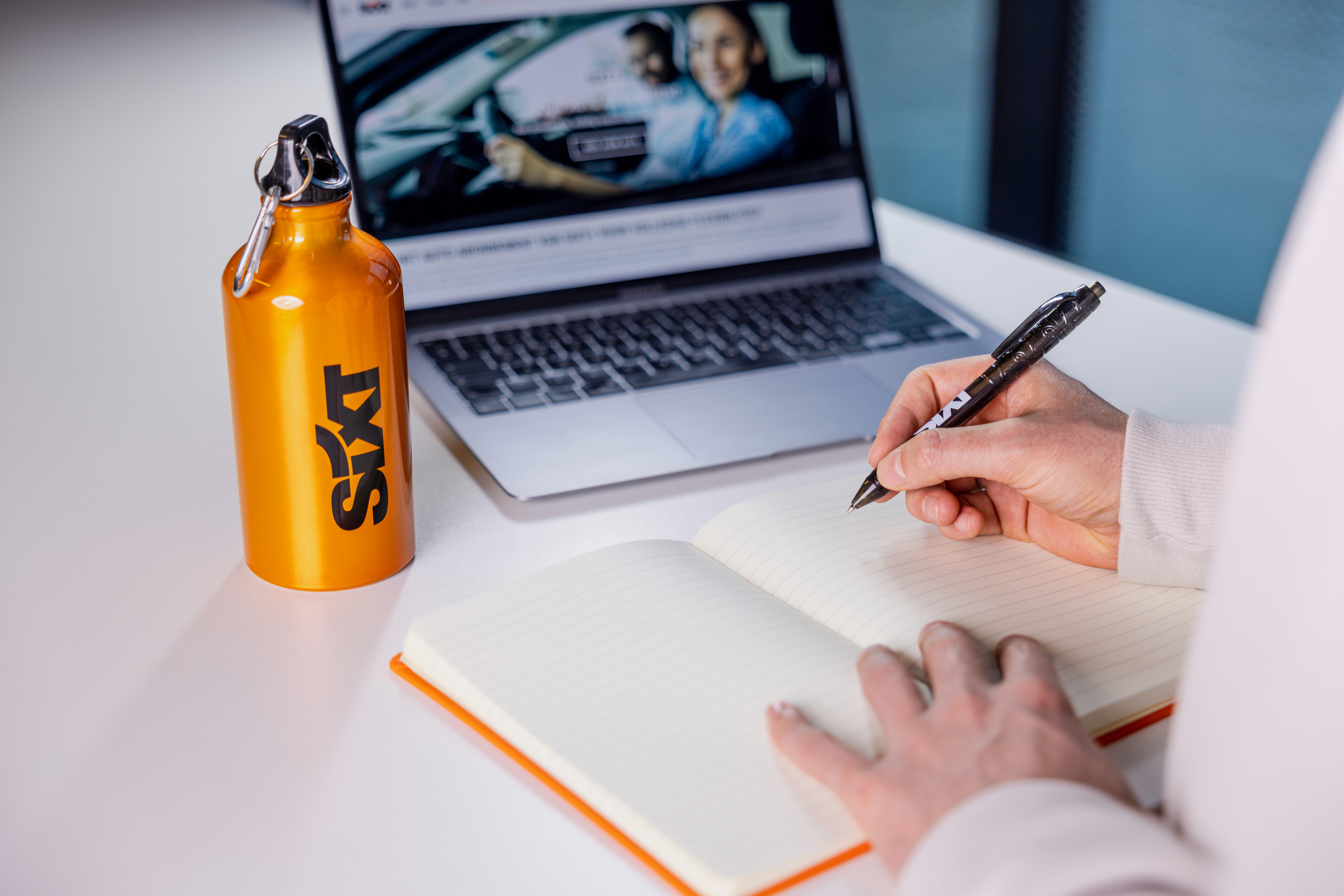 Une personne écrivant dans un carnet à côté d'une bouteille d'eau orange "Sixt" et d'un ordinateur portable ouvert sur une table