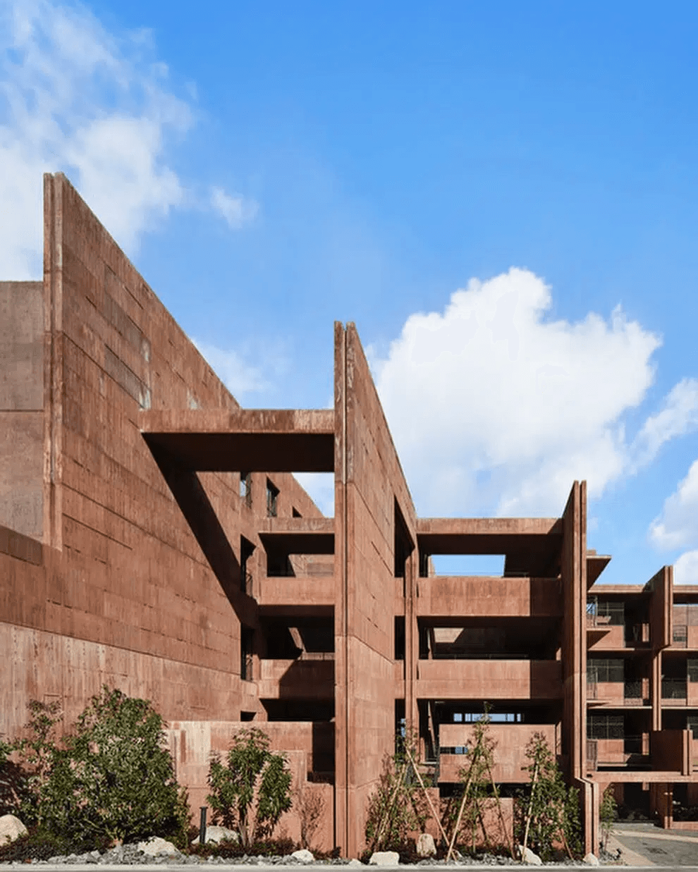 Contemporary brutalist building with tall reddish-brown concrete walls, elevated connecting walkways, and geometric openings beneath a bright blue sky.