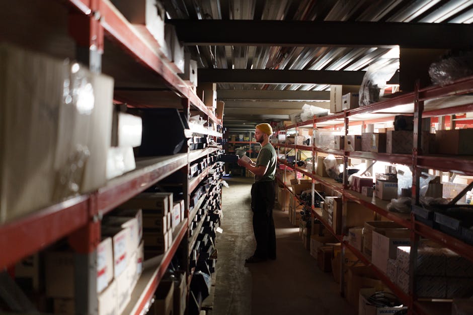 A warehouse worker standing between two rows of red metal shelving units stocked with boxes, bins, and packaged goods, reviewing inventory on a clipboard in a dimly lit storage facility.